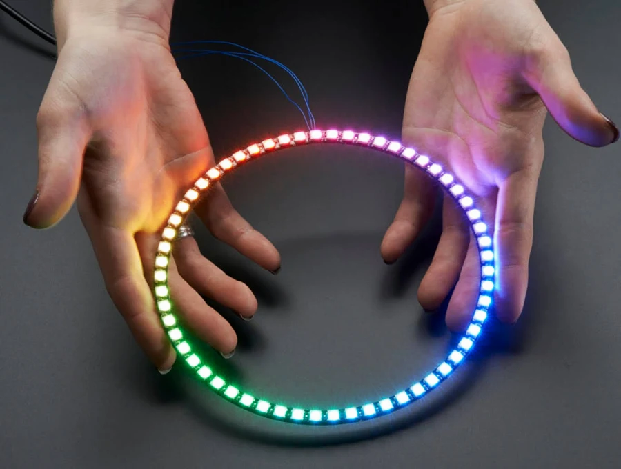 Two hands are holding a circular LED light ring that emits colorful lights. The ring has multiple colored lights along its edge, transitioning through hues of red, green, and blue. It is set against a dark background, and the connection wires are visible, extending from the top of the ring.
