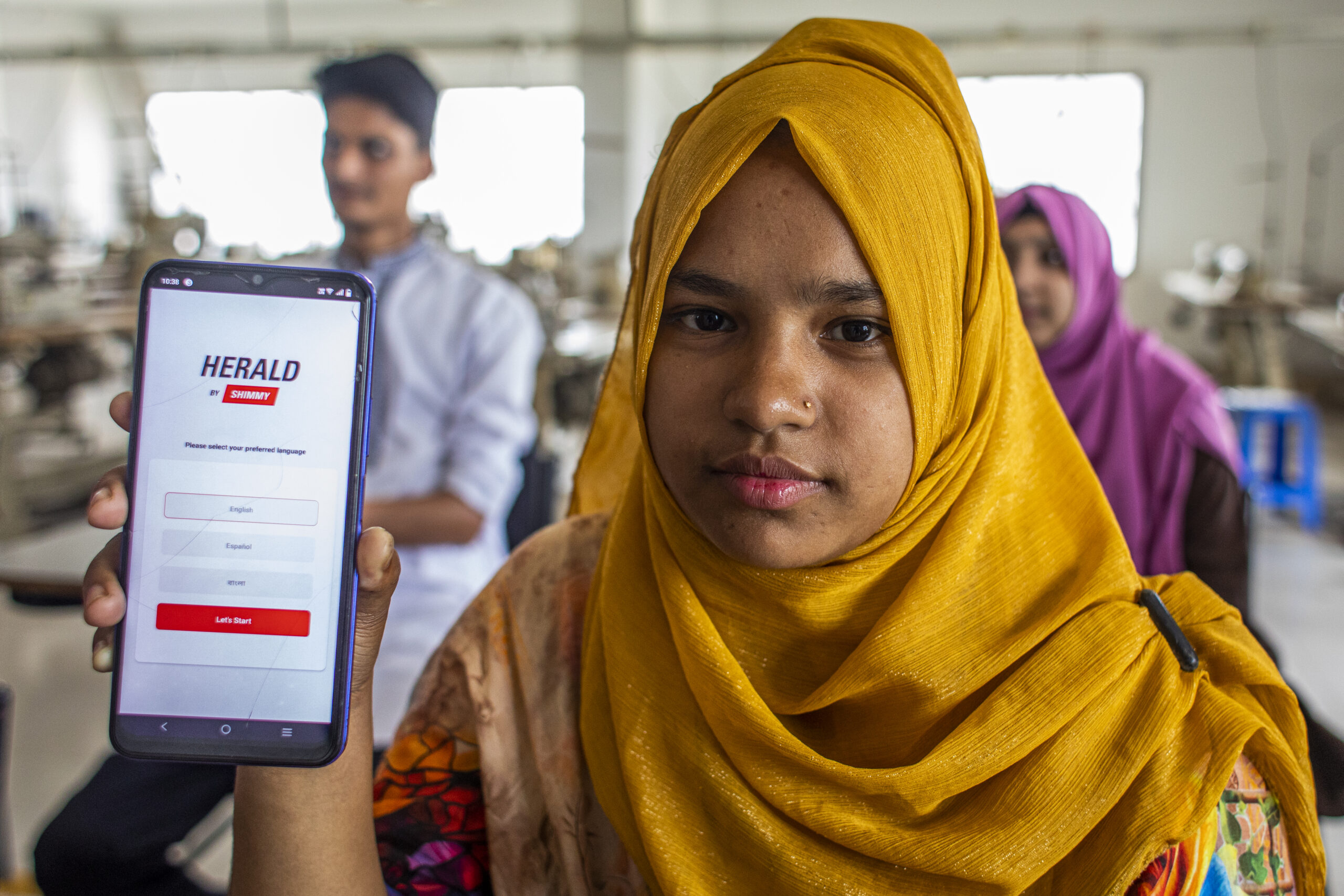 A young woman wearing a bright yellow hijab holds up a smartphone displaying the HERALD app interface, which prompts users to select a preferred language. The setting appears to be a workshop with sewing machines.
