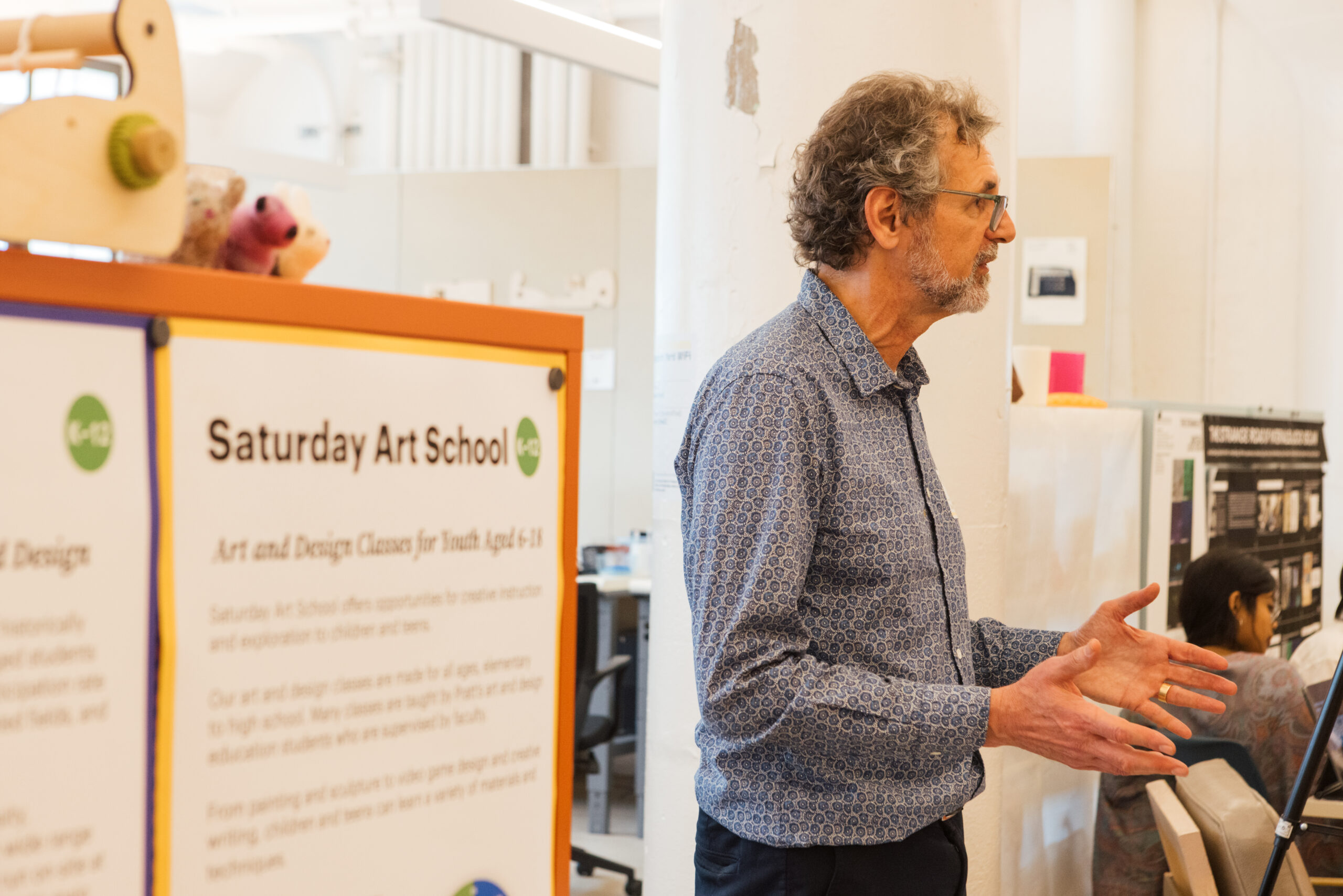 A man with curly hair and glasses stands beside an orange display board labeled "Saturday Art School." He gestures with his hands while speaking. The background shows other people working at desks and various art-related materials. The display board provides information about art and design classes for youth aged 4-18.