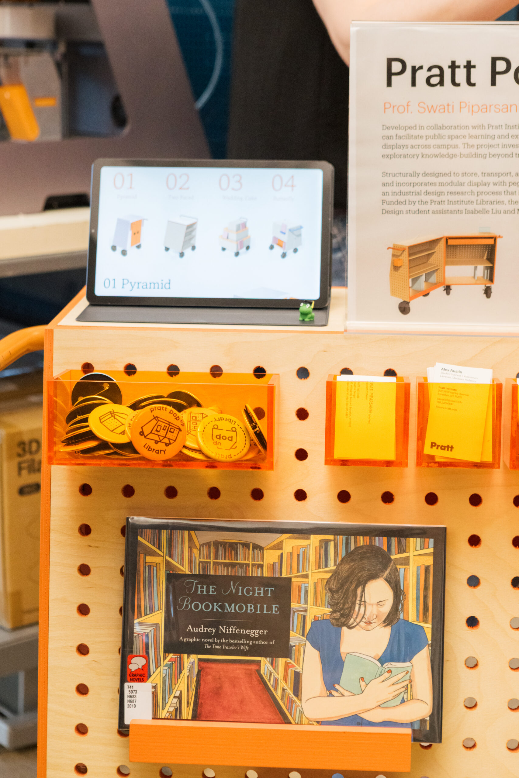 A display showcasing a tablet and wooden cart at a library. The tablet lists four designs: Pyramid, Two Placed, Modeling Cart, and Assembly. Below the tablet, there are orange boxes containing round tokens with library-themed designs. A book titled "The Night Bookmobile" by Audrey Niffenegger is prominently featured at the bottom, displaying an illustration of a woman reading in a library.