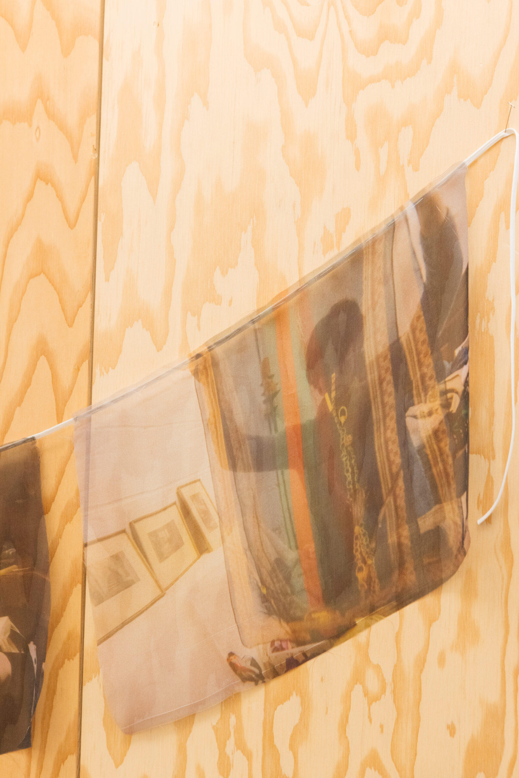 A sheer fabric banner is displayed against a wooden wall. The banner features a collage of images, including partially visible photographs exhibited on a wall. The overall effect creates a translucent overlay of colors and shapes, blending the images together. The background consists of light wood grain patterns, enhancing the texture of the setting.