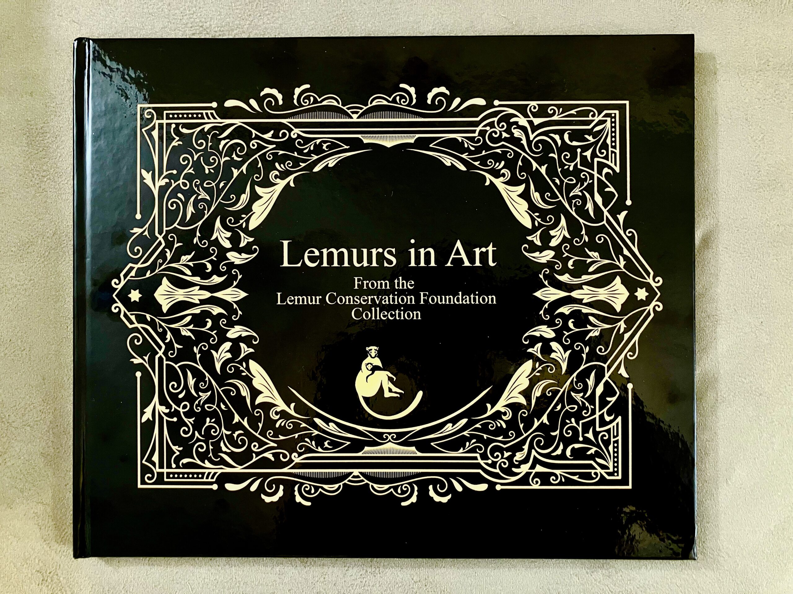 A black book cover titled "Lemurs in Art" is centrally displayed, with decorative white floral and scrollwork around the edges. Below the title, it notes that the book is from the Lemur Conservation Foundation Collection. The glossy finish reflects light, enhancing the ornate design.