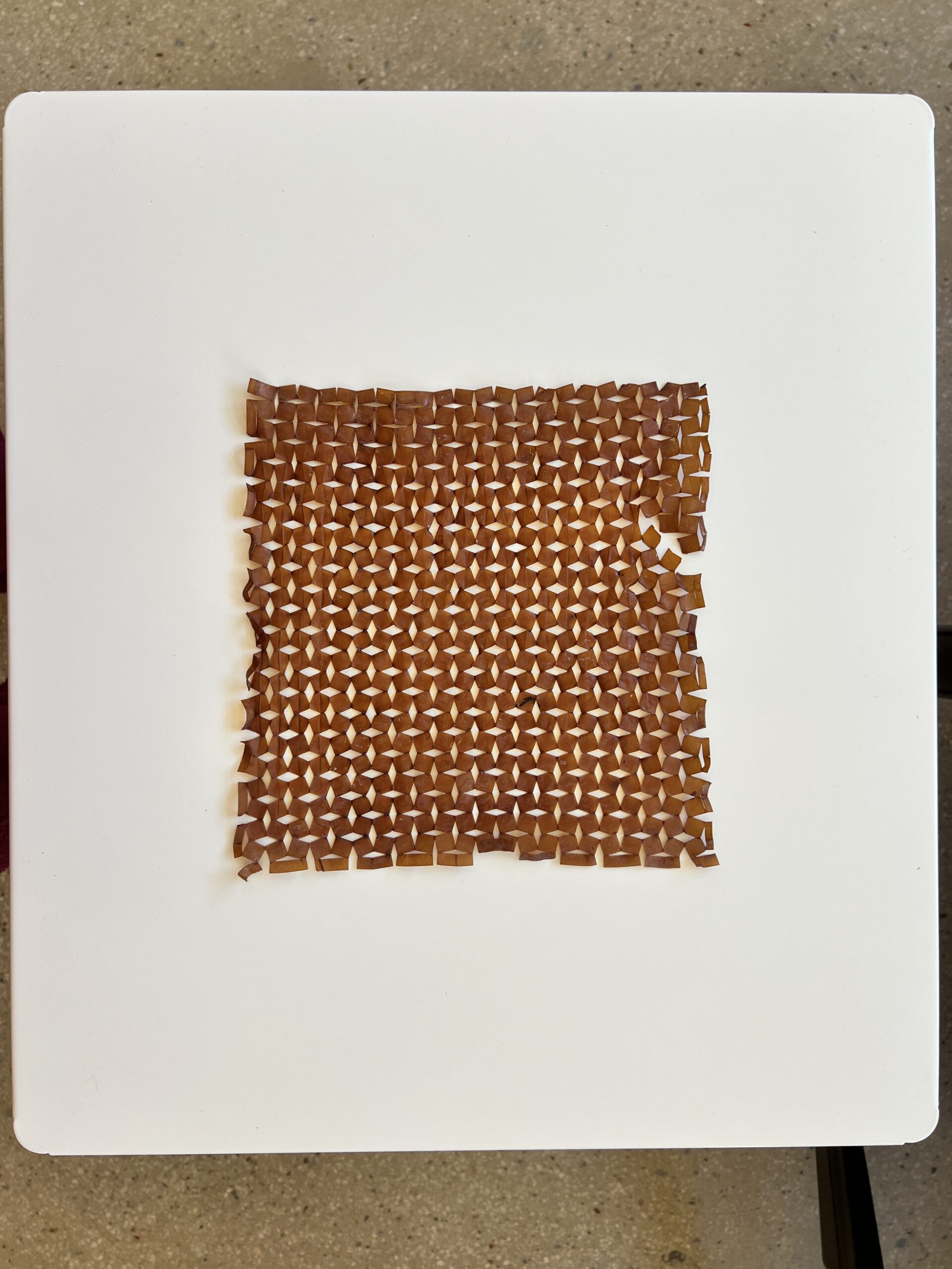 A square mat is composed of small brown, triangular-shaped tiles arranged in a woven pattern. The tiles create a textured surface with varying shades of brown, placed on a white table with a smooth finish. The edges of the mat appear slightly irregular. The background shows a gray surface underneath the table.