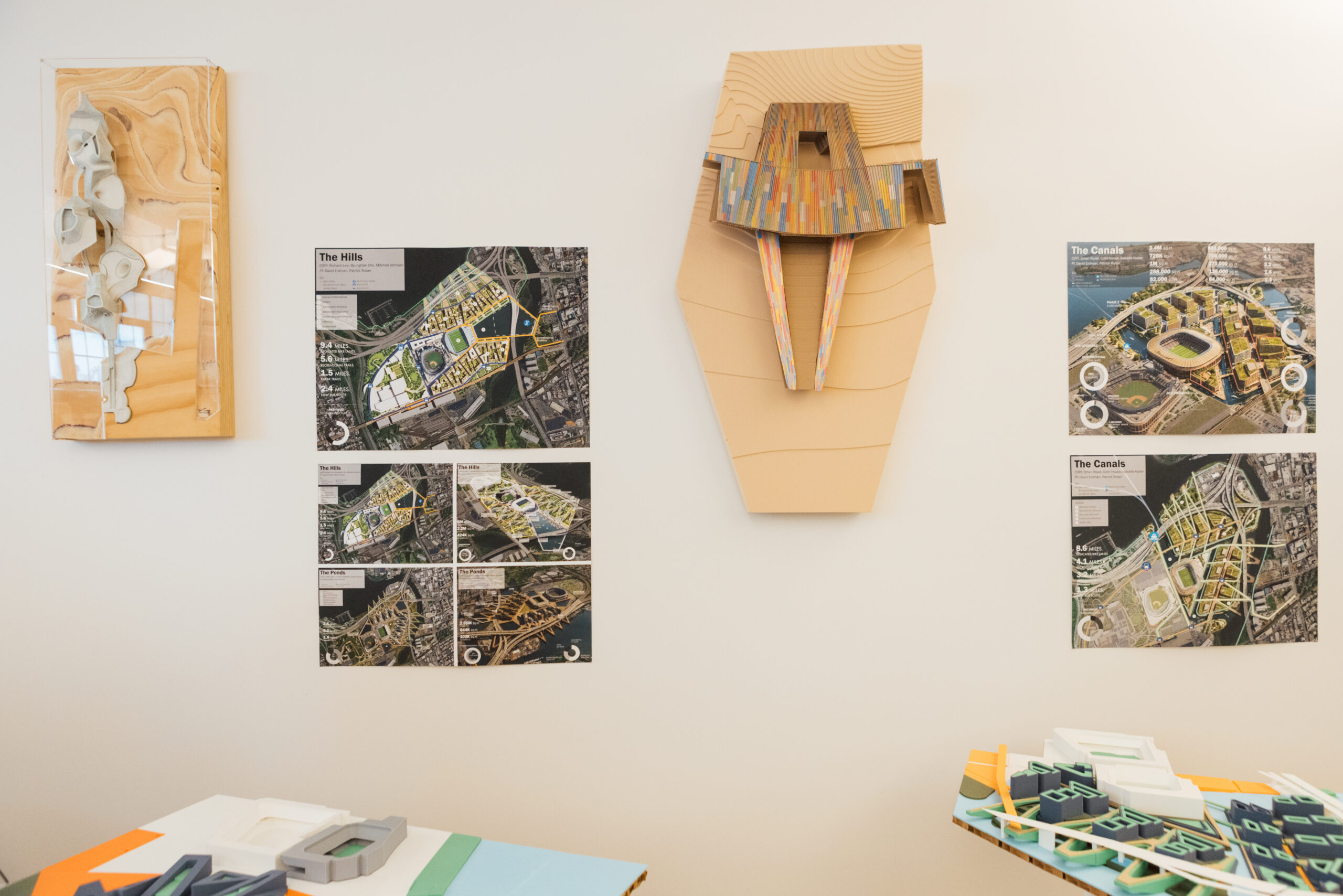 A display featuring architectural models and design plans. On the left, a 3D model in clear casing with beige layers. In the center, a large, colorful model mounted on the wall resembling a structure with extending parts. Surrounding it are posters detailing plans for "The Hills" and "The Canals," showing aerial views of urban designs with descriptions. A table below contains various colorful architectural model parts.