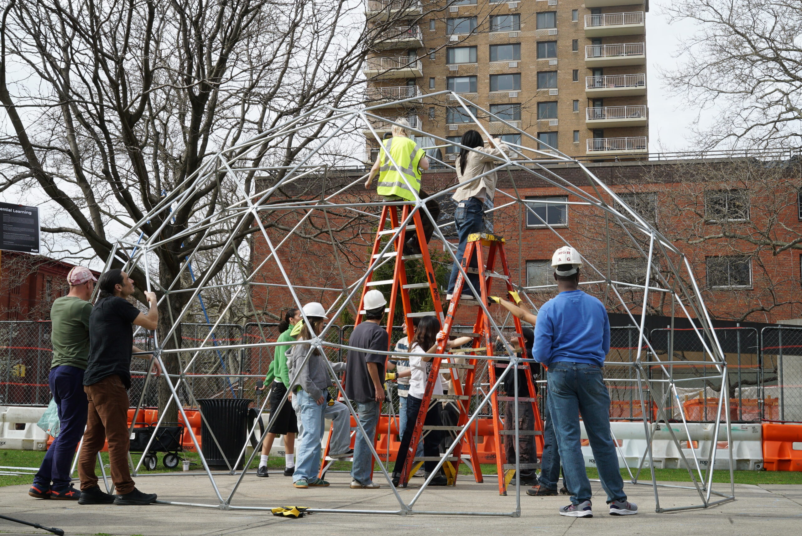 A group of nine people is assembling a large geodesic dome structure in an outdoor setting. Some individuals are on ladders attaching parts, while others are on the ground holding components. Nearby, there are trees without leaves, and a multi-story building in the background. Construction signs and safety barriers are also visible in the area.