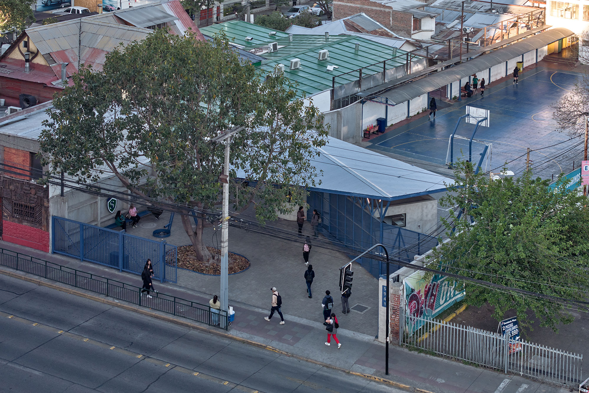 An aerial view of a neighborhood featuring a sports facility with blue courts and a distinct roof shape. Several people are walking along the sidewalk and entering the facility, while a large tree provides shade nearby. In the background, various rooftops and structures are visible, along with a streetlight and some urban graffiti on the wall to the right.