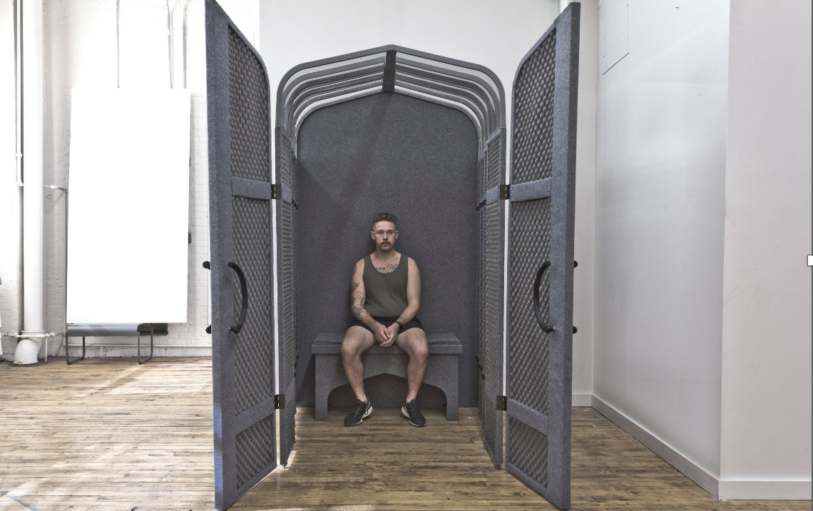 A man with a tattoo sits on a bench inside a large, open structure with textured walls that create a somewhat enclosed space. The structure has two doors on either side and is located in a bright room with wooden flooring. Natural light streams in from large windows.