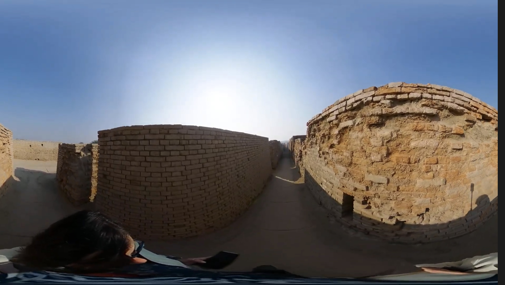 A wide pathway runs between tall, ancient brick walls, which display various shades of brown and gray. The sun shines brightly from above, illuminating the scene. In the foreground, part of an individual with dark hair can be seen, holding a device. The setting suggests an archaeological site or historical ruins in a desert-like environment.