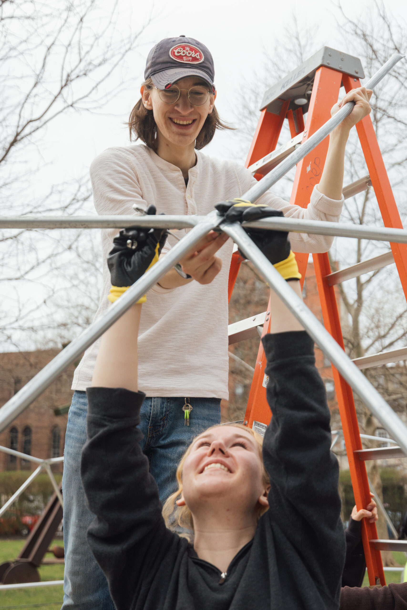 Two individuals are collaborating to assemble a metal structure outdoors. One person, standing on a ladder, is smiling and holding a pipe, while another person, underneath, raises their arms with a joyful expression, gripping a lower section of the structure. Both are wearing gloves, and trees and a building are visible in the background.