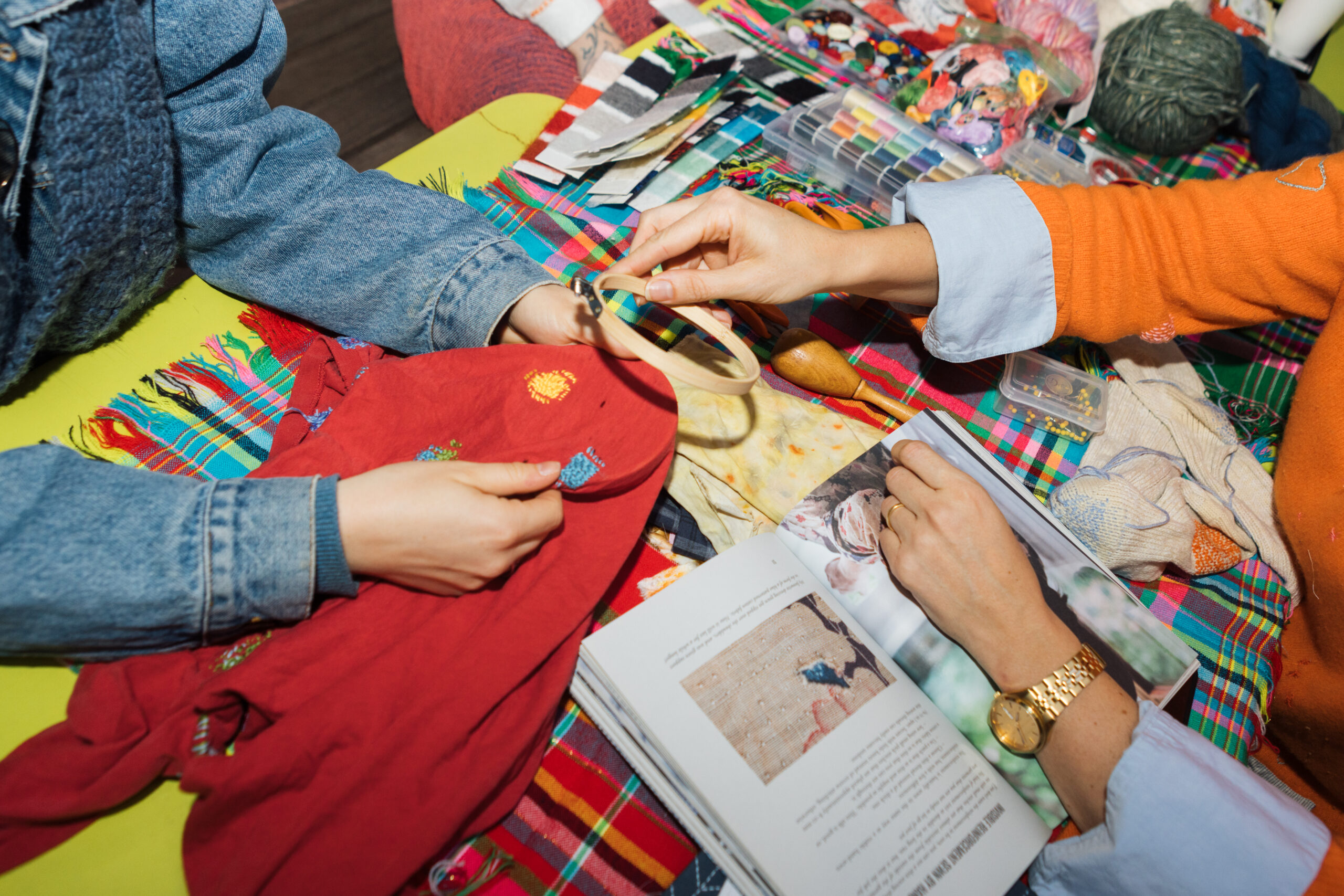 Two people are engaged in a creative activity on a colorful, patterned table. One holds a red garment featuring embroidery, while the other is grasping an embroidery hoop. A book lies open with images and text, and various craft materials, such as threads and fabric swatches, are scattered across the table, illustrating a vibrant crafting environment.