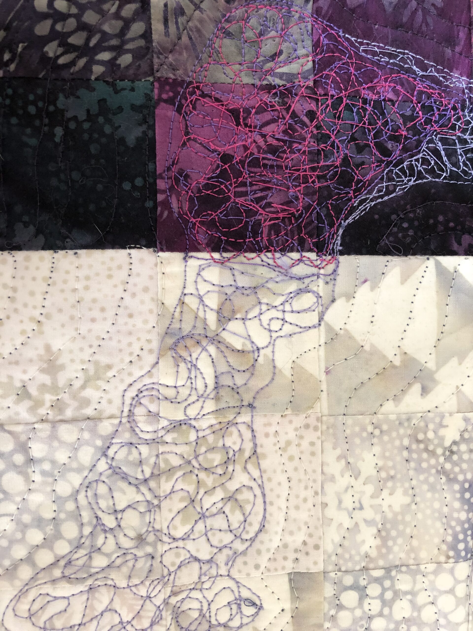 A close-up image of a quilt featuring various patches in shades of purple, black, and cream. The top left area has a dark green patch with texture, while the top right showcases intricate stitched patterns in pink and purple. The bottom half comprises light-colored patches with varying patterns and subtle stitching. The overall design combines geometric shapes with freeform embroidery, creating a blend of color and texture.