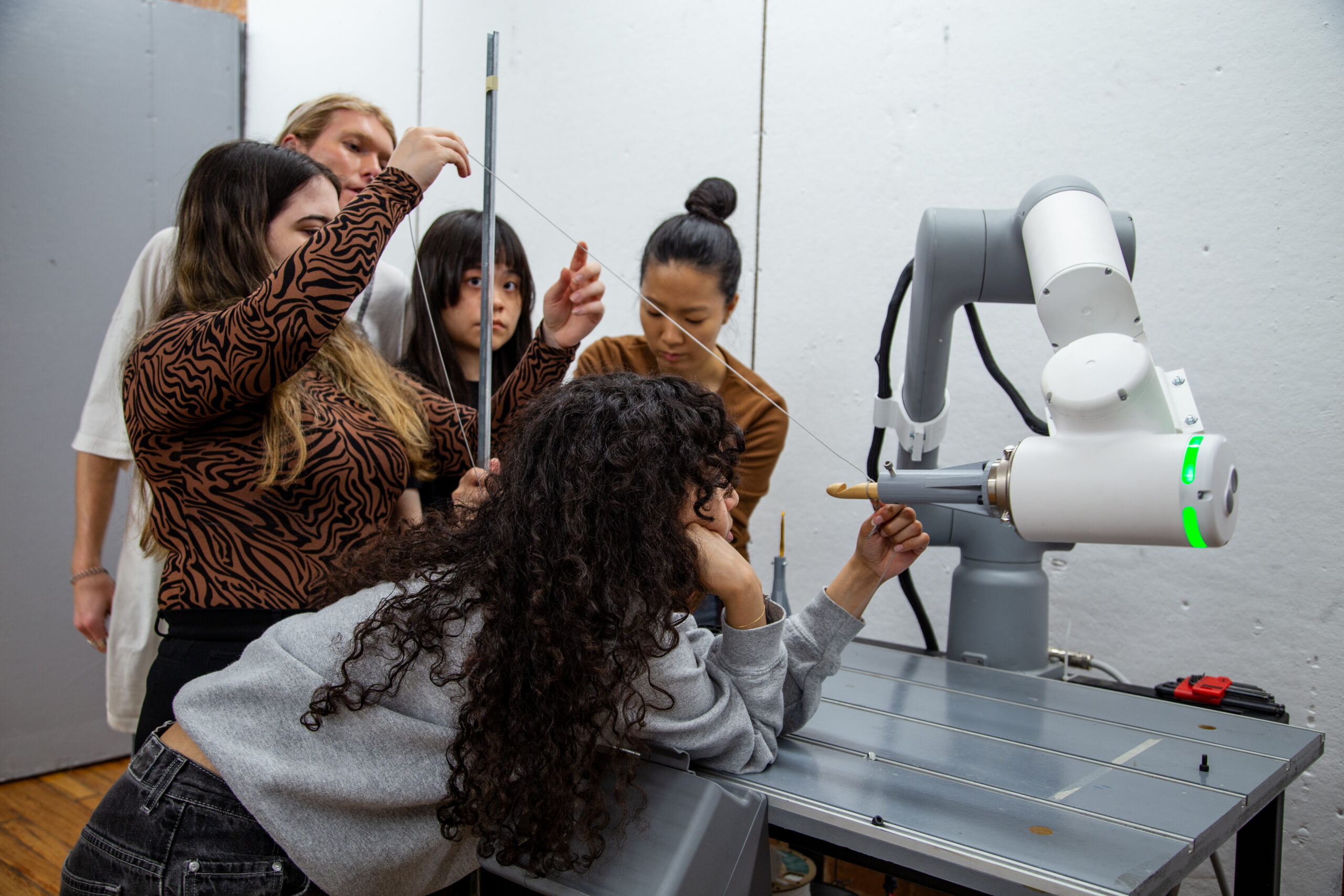 A group of five individuals is gathered around a robotic arm in a workshop setting. One person, leaning on the table, appears to be guiding or observing a string attached to the robotic arm, while another is holding a vertical metal rod. The others are focused on the task, contributing to a collaborative project involving technology and design. The workspace has a minimalist aesthetic with a simple background.