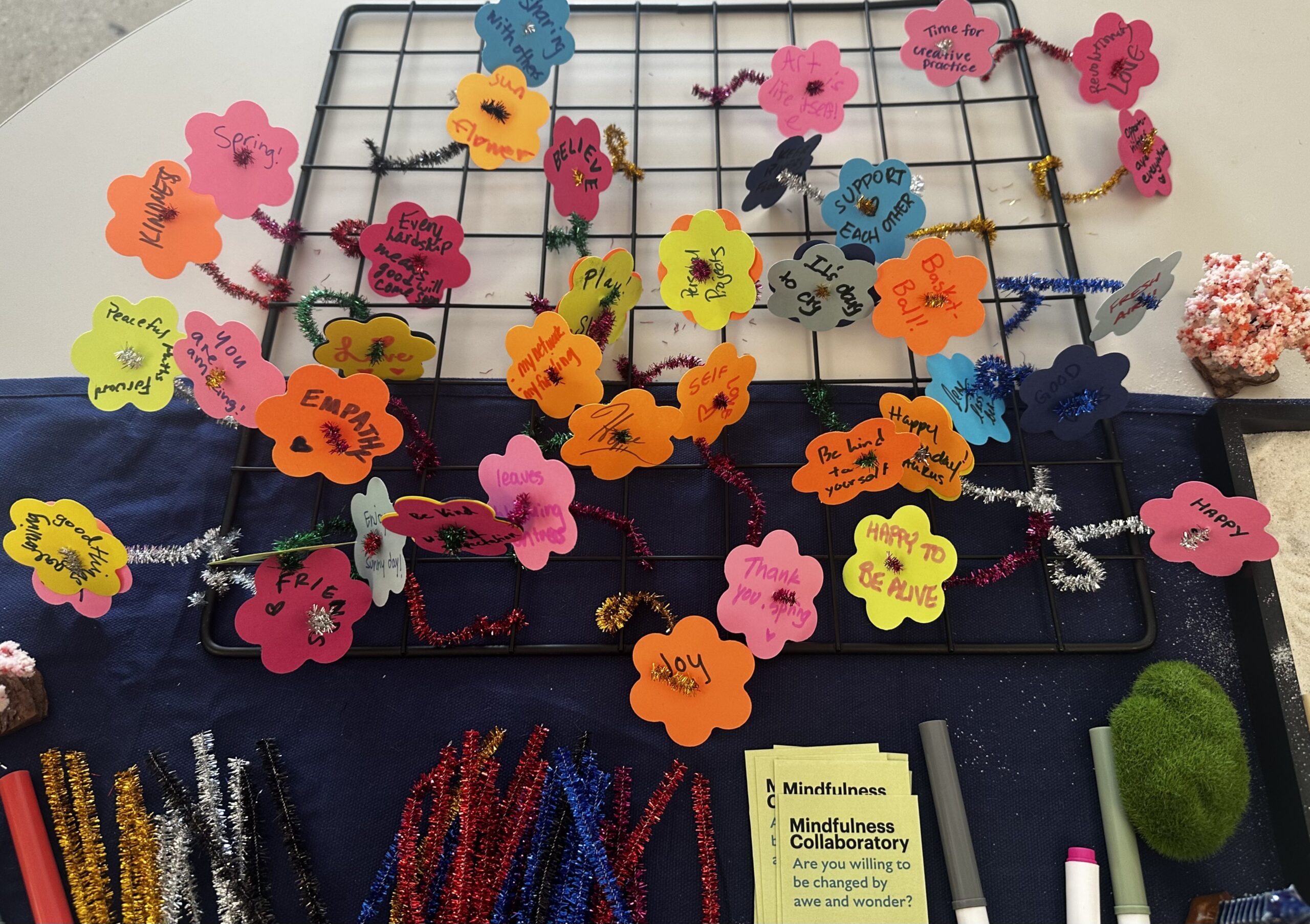 A wire grid displays colorful flower-shaped notes with handwritten words and phrases, such as "Believe," "Empathy," and "Happy to be alive." Note connectors made of pipe cleaners intertwine the notes. Below the grid, various colored pipe cleaners are spread out alongside a green decorative item and markers. Yellow pamphlets titled "Mindfulness Collaboratory" are also visible, encouraging engagement in mindfulness activities.