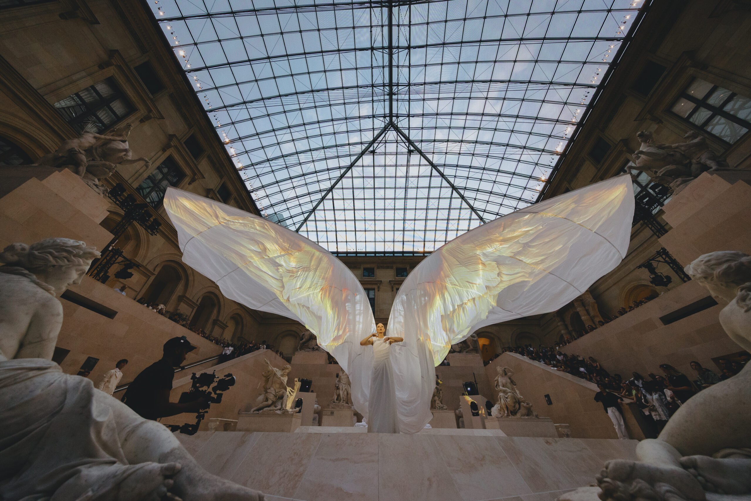 In a glass-ceilinged atrium in the Louvre, a person stands wearing a gown and giant fabric wings. Onto the wing-shaped form of the plain fabric is projected a detailed image of wings.