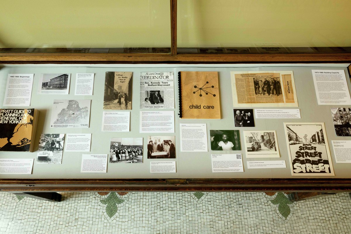 A display case showcasing historical materials related to community planning and childcare. The collection includes photographs, newspaper articles, and pamphlets, arranged on a flat surface. Featured items include a booklet titled "Pratt Guide to Planning and Renewal for New Yorkers," various images from community events, and an article about a tour led by Sen. Kennedy. The background shows patterned tile flooring.