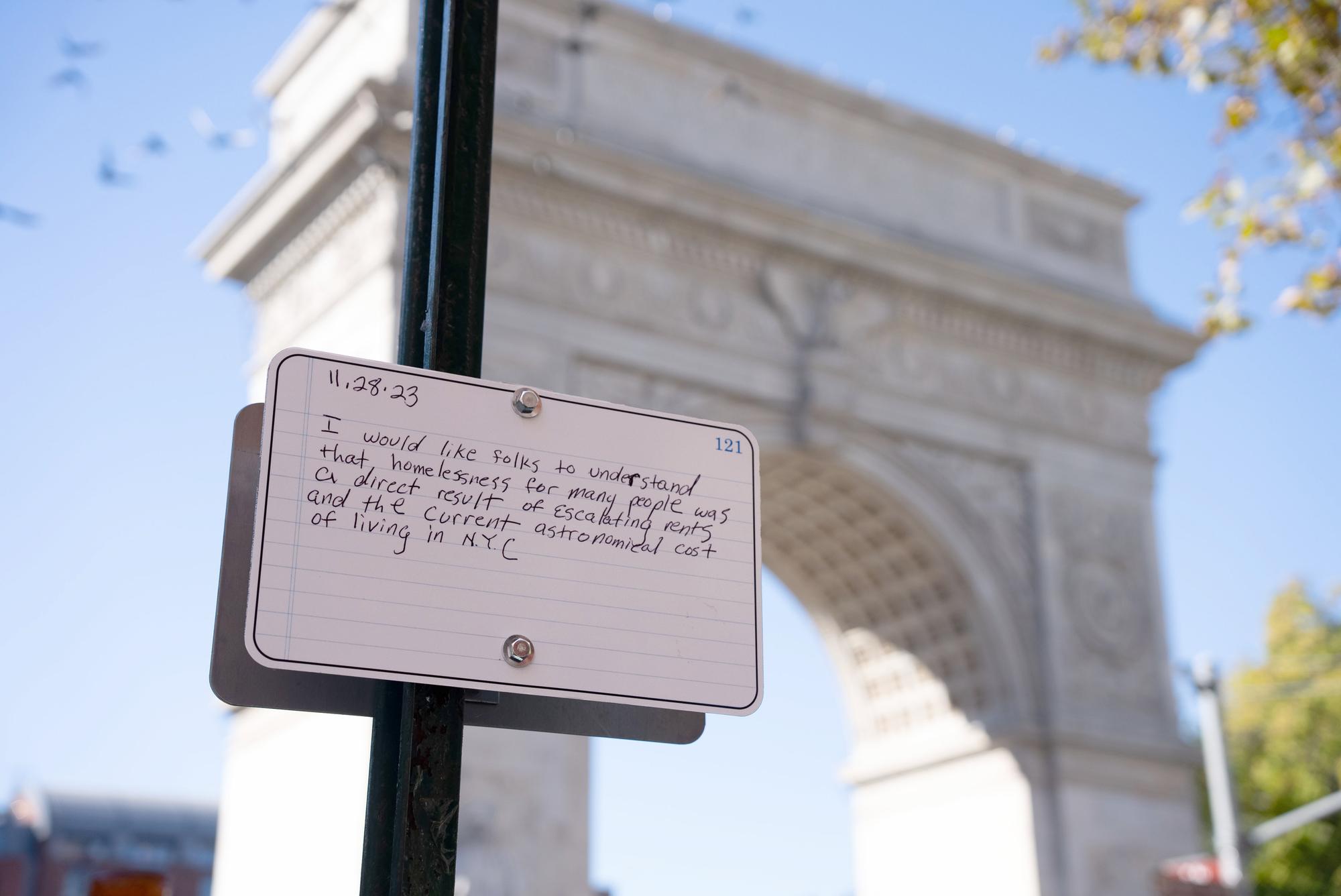 A handwritten note is attached to a pole in front of a stone archway, with a clear blue sky in the background. The note reads: "11.28.23 I would like folks to understand that homelessness for many people was a direct result of escalating rents and the current astronomical cost of living in N.Y.C." Birds can be seen in the sky around the archway.