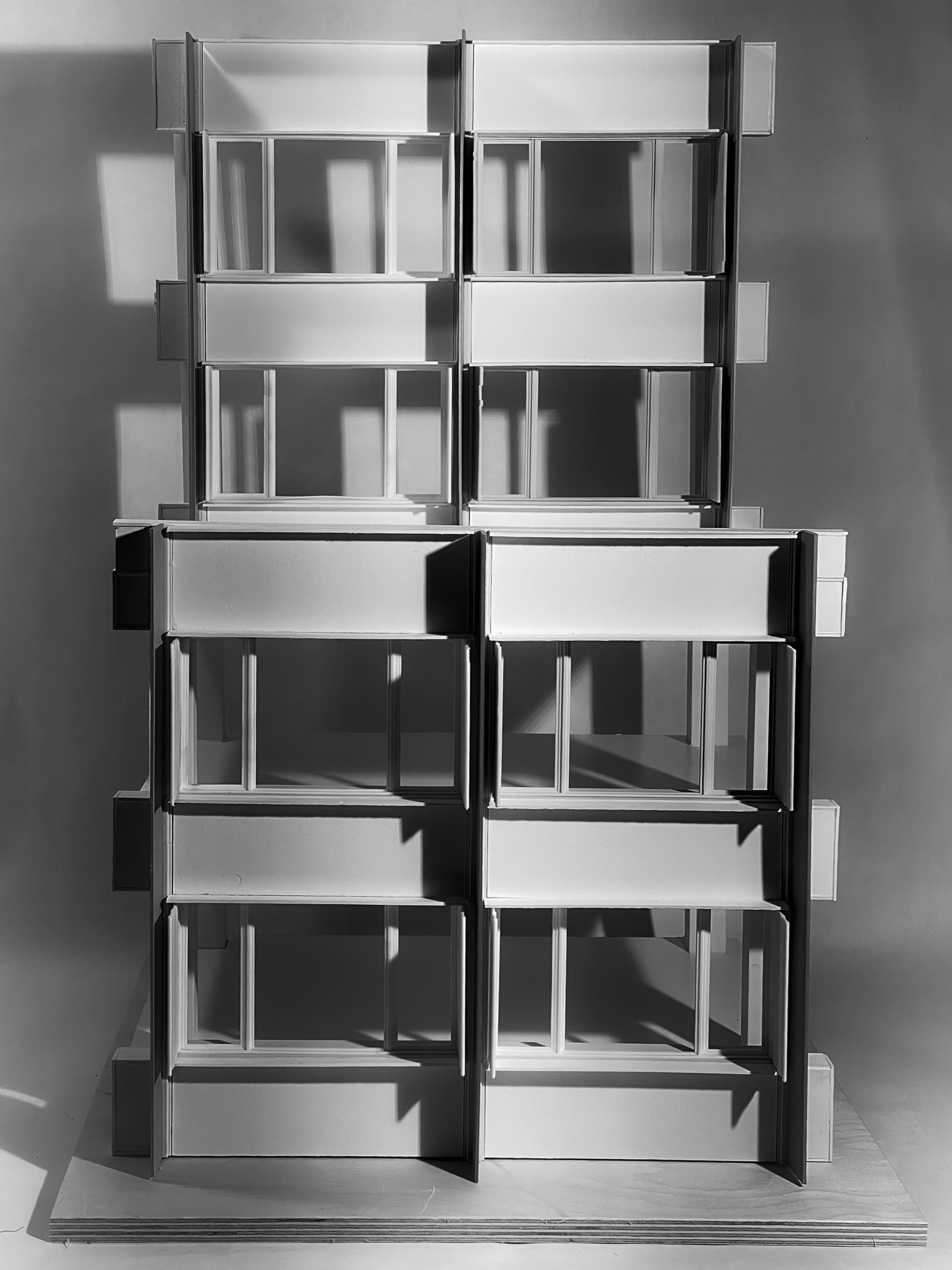 A monochromatic model of a building or structure with multiple levels. The design features several modular sections, each with rectangular windows and a flat rooftop. Shadows cast by the structure enhance the geometric details, while the overall composition presents a clean, minimalistic aesthetic against a light background.