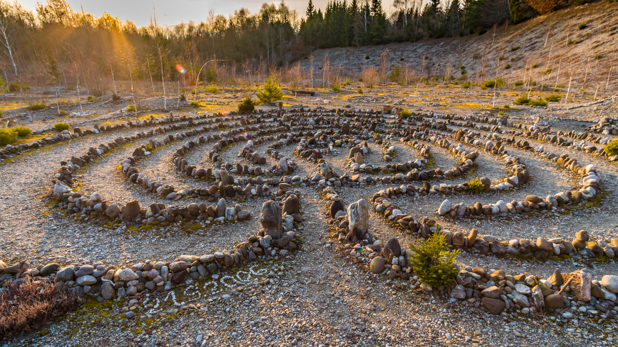 A stone labyrinth is set in a natural landscape, with pathways outlined by rocks of various sizes. The sun is setting in the background, casting a warm glow over the area, which features sparse vegetation and patches of gravel. The labyrinth's design is circular with winding paths, and small trees are scattered around the edges.