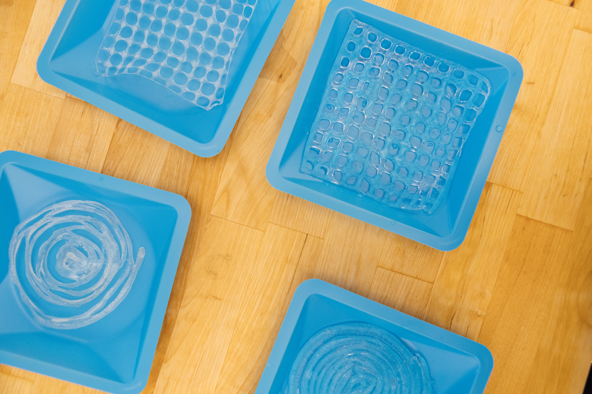 Four blue silicone molds placed on a wooden surface, each containing different gelatin shapes. One mold has a flat, textured sheet resembling bubble wrap, another is a circular spiral, and the rest feature unique geometric designs.