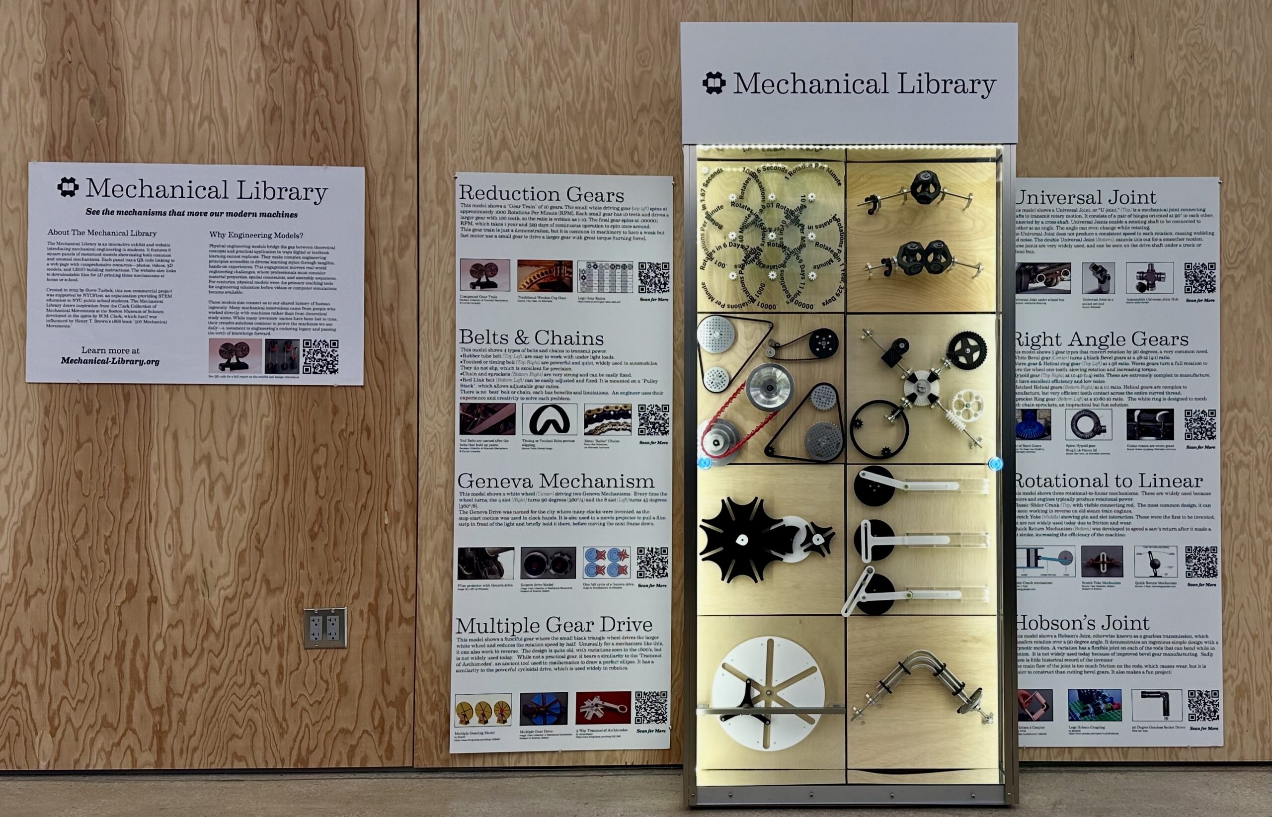 A display labeled "Mechanical Library" showcases various mechanical models, including gears, chains, and joints. The back wall features informative posters about different mechanisms, such as Reduction Gears, Universal Joints, and the Geneva Mechanism. Each poster contains descriptions and QR codes for further learning. The wooden paneling adds a natural aesthetic to the exhibit, with well-organized sections for each topic. Several mechanical components are highlighted in a lit cabinet.