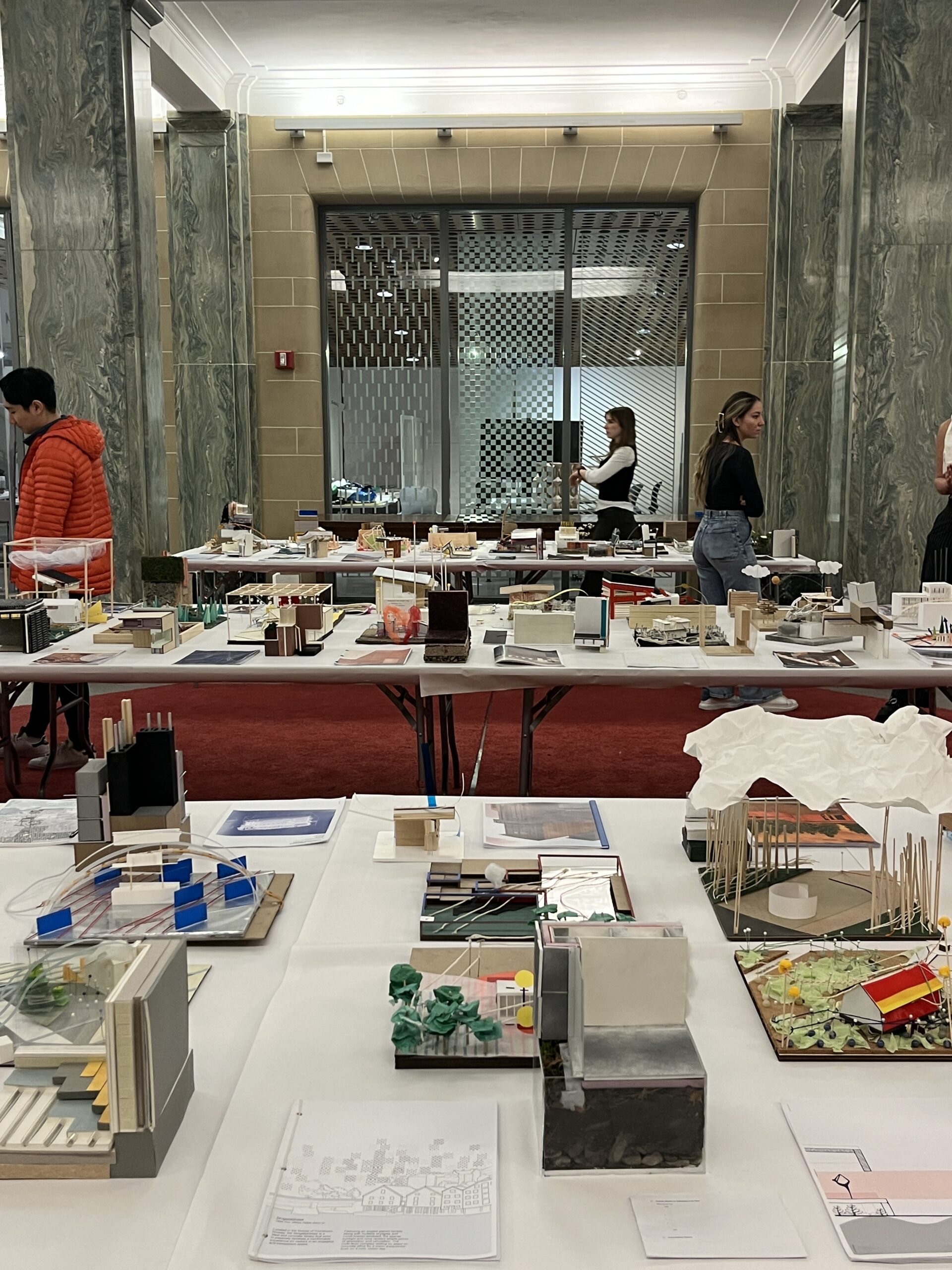 A spacious exhibition area displays multiple architectural models and drawings on tables. Attendees are engaged, with two individuals visible in the foreground and others observing or discussing the models. The backdrop showcases large windows with a patterned façade, while the floor is covered in red carpeting. Various model styles, including some with greenery and structures, are arranged across the tables.