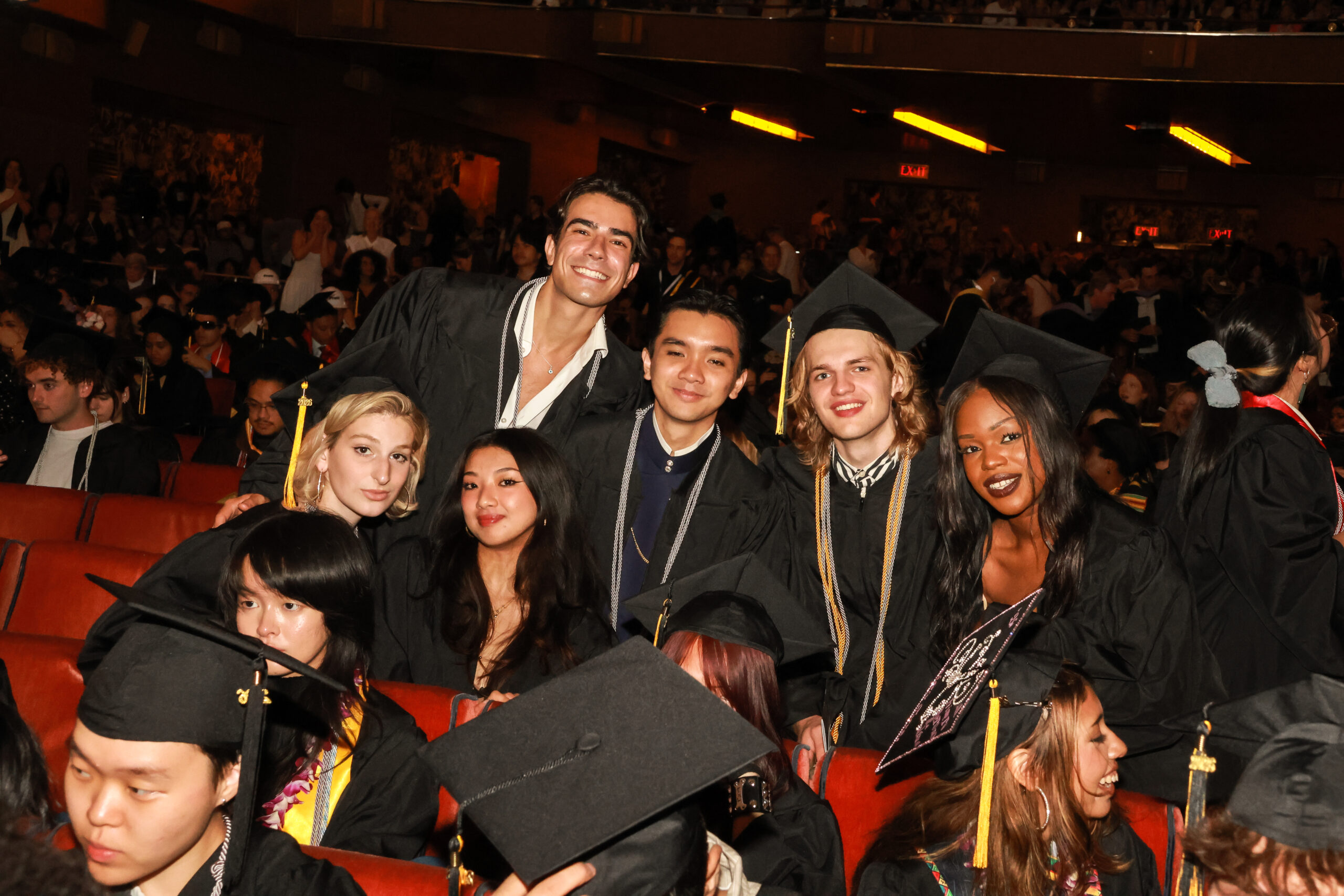 A group of six graduates, wearing black caps and gowns, pose together at a graduation ceremony. They are seated in a crowded auditorium with red seats. Each graduate is smiling, and some are leaning towards one another for a closer group photo. The background shows other attendees, adding to the celebratory atmosphere of the event.