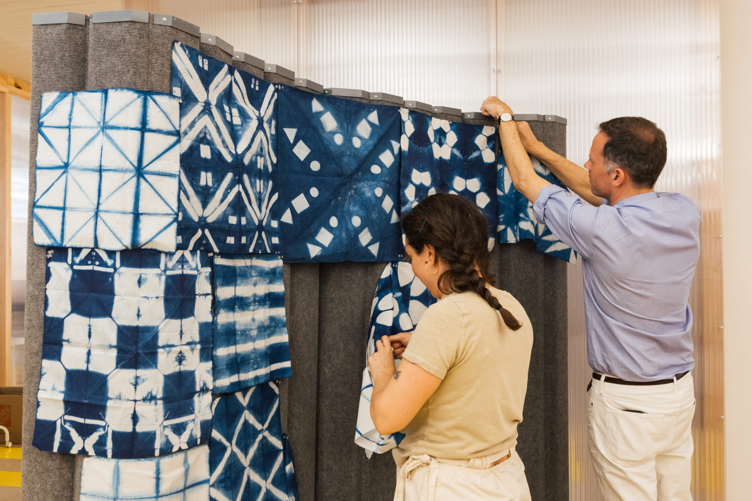 Two individuals are arranging indigo-dyed textile patterns on a display board. One person, wearing a light-colored shirt, is placing a fabric square on the wall, while the other, in a blue button-up shirt, is hanging fabric at the top. Various patterned textiles in shades of blue and white are visible, showcasing different designs and textures. The setting appears bright and creative, likely in a studio or workshop environment.