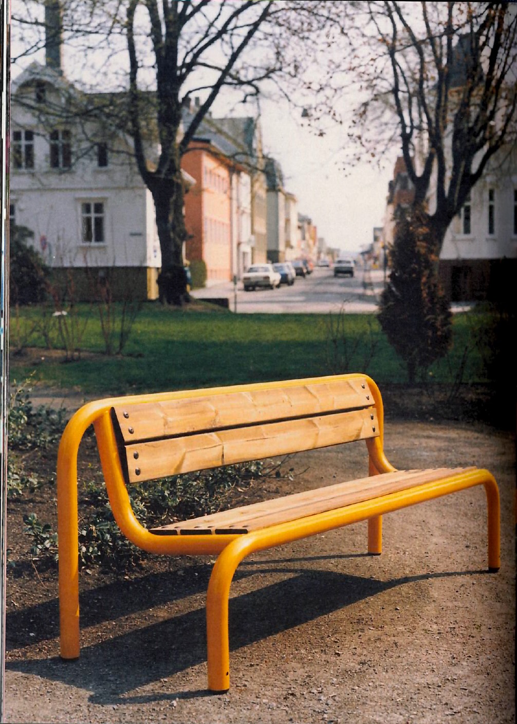 A bright yellow, modern-style bench with a wooden seat is positioned on a gravel path. In the background, trees and buildings line a quiet street, with a few parked cars visible. The scene is illuminated by natural light, suggesting a pleasant day.