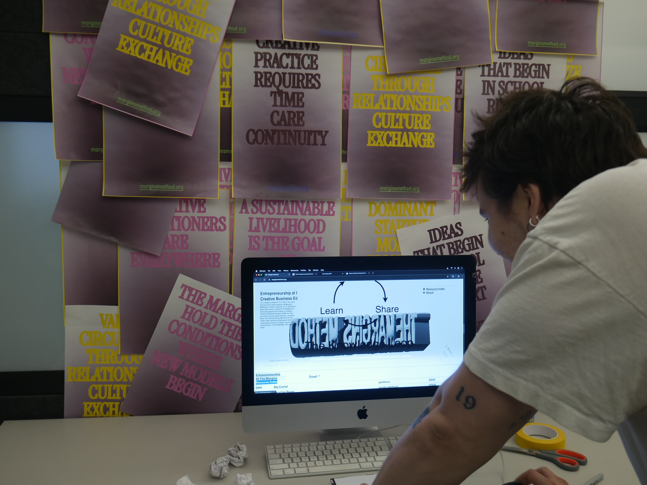 A person is leaning over a computer monitor displaying a webpage with the text "Learn" and "Share" on it. Behind them is a wall covered with colorful posters featuring phrases related to relationships and creative practice. The workspace includes crumpled paper and tape, suggesting an active creative environment. The individual has a tattoo on their arm and is wearing a light-colored shirt.