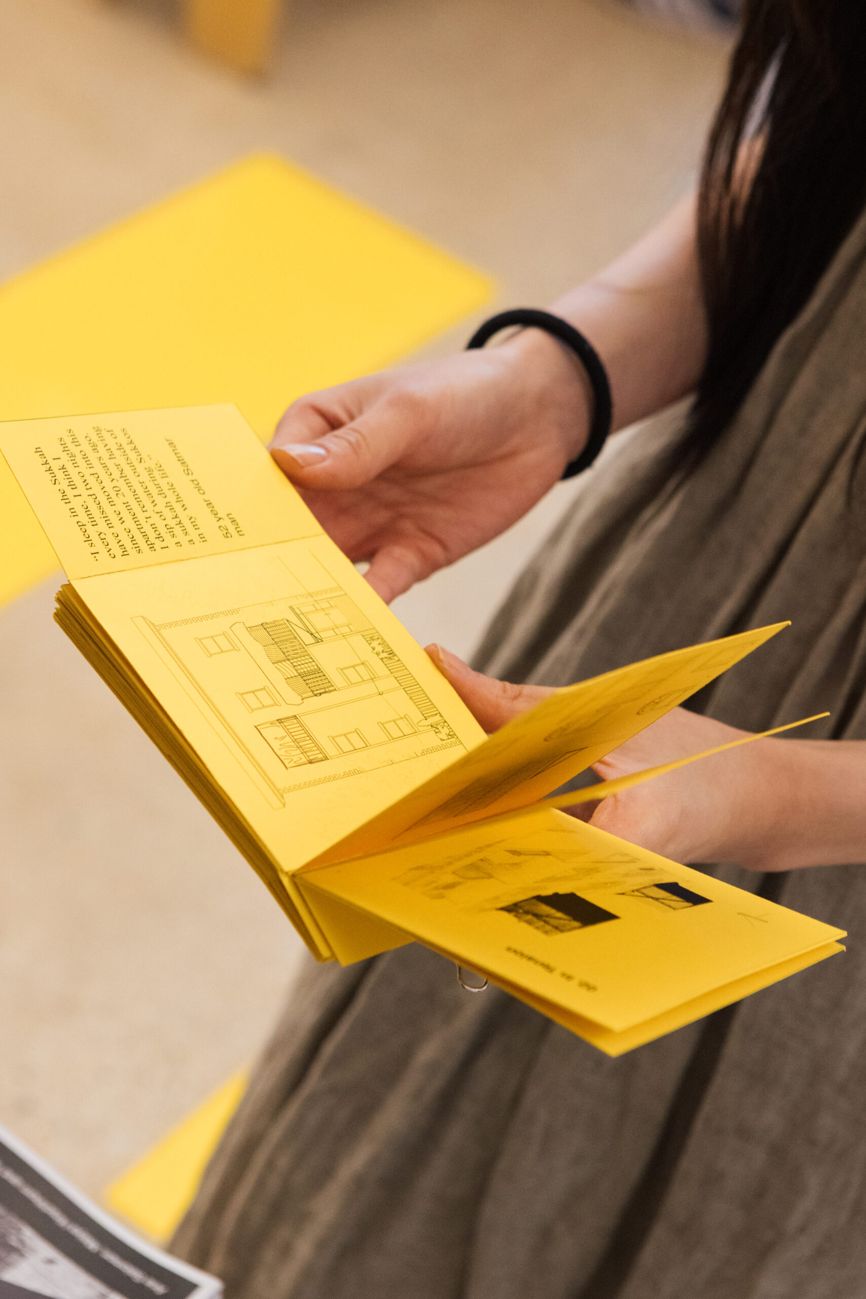 A person is holding an open yellow booklet with illustrations and text. The visible page features a drawing of a building, accompanied by descriptive text. The background includes a hint of other yellow surfaces, suggesting a vibrant environment. The person's attire is casual, with their hand prominently showing the booklet.