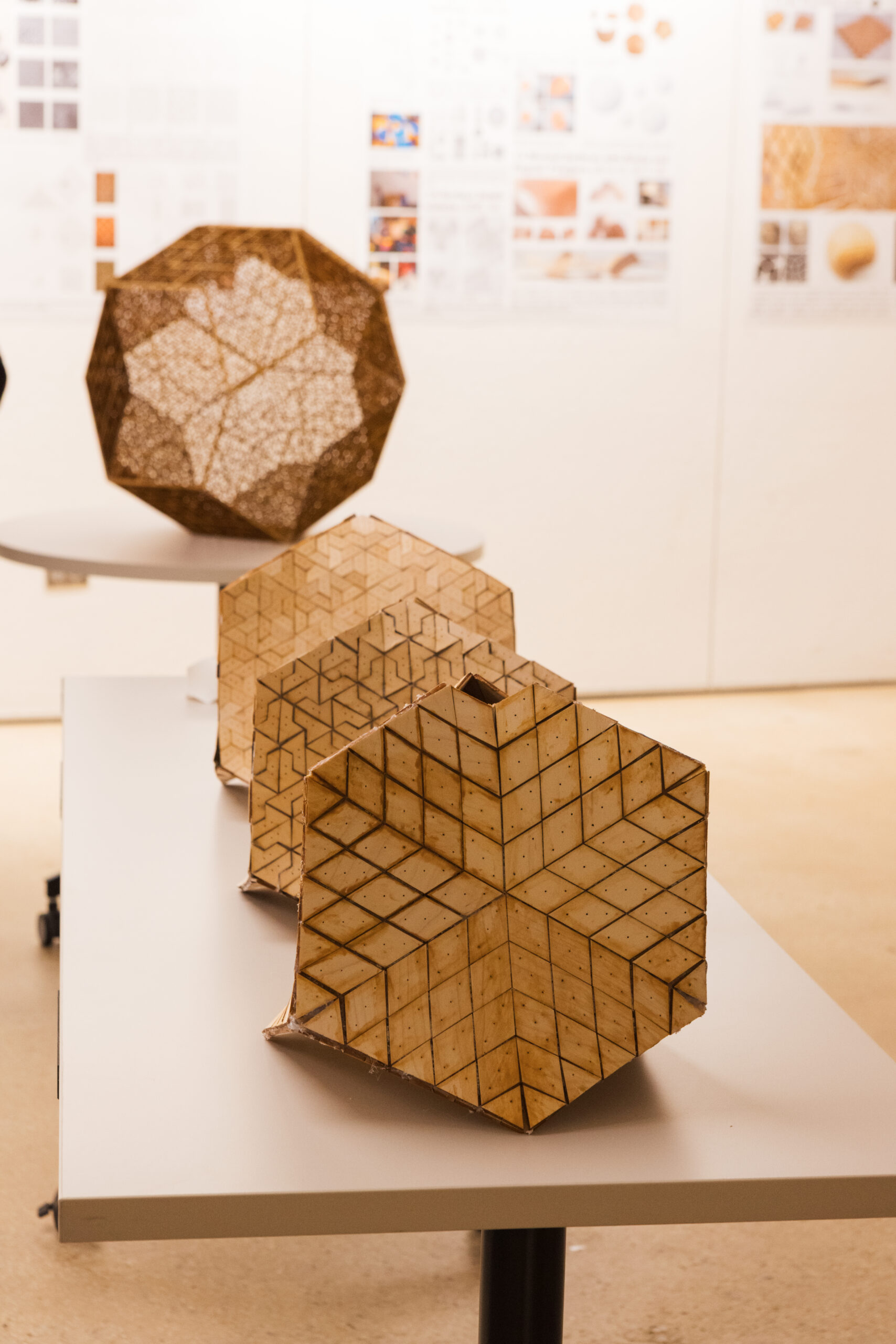 Three geometric wooden sculptures are displayed on a white table. The sculptures feature intricate patterns and cutouts, showcasing different shapes, including a hexagonal form in the foreground and more rounded shapes at the back. In the background, posters with various designs and patterns are partially visible, adding context to the artistic setting.