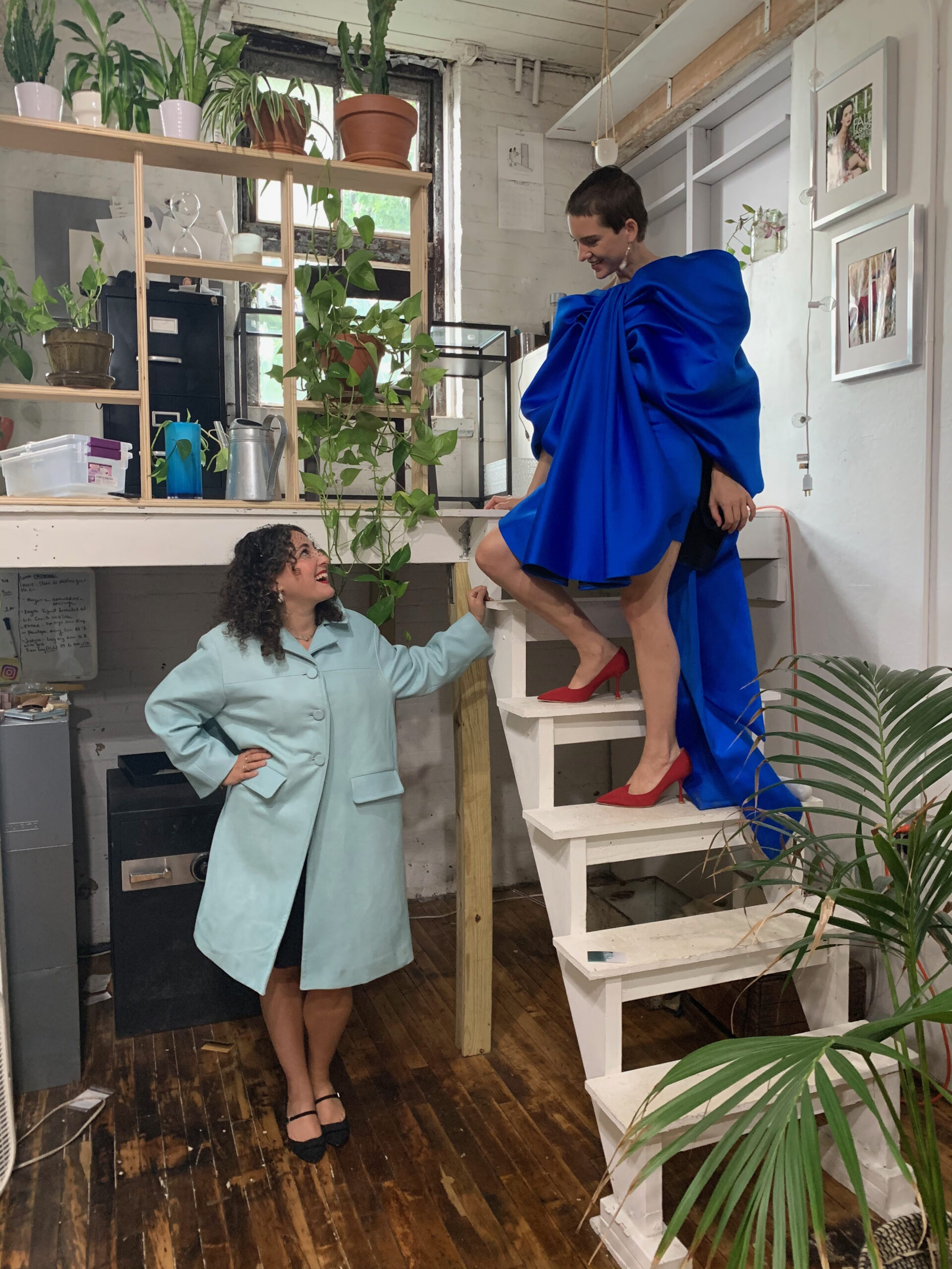 Two individuals pose in a stylish, creative space. One person is sitting on a staircase, wearing a voluminous blue dress and red heels, while the other stands on the lower step, wearing a light blue coat and black shoes. They share a joyful interaction amidst plants and decor, with shelves in the background displaying various items. The floor is wooden, and the atmosphere feels vibrant and artistic.