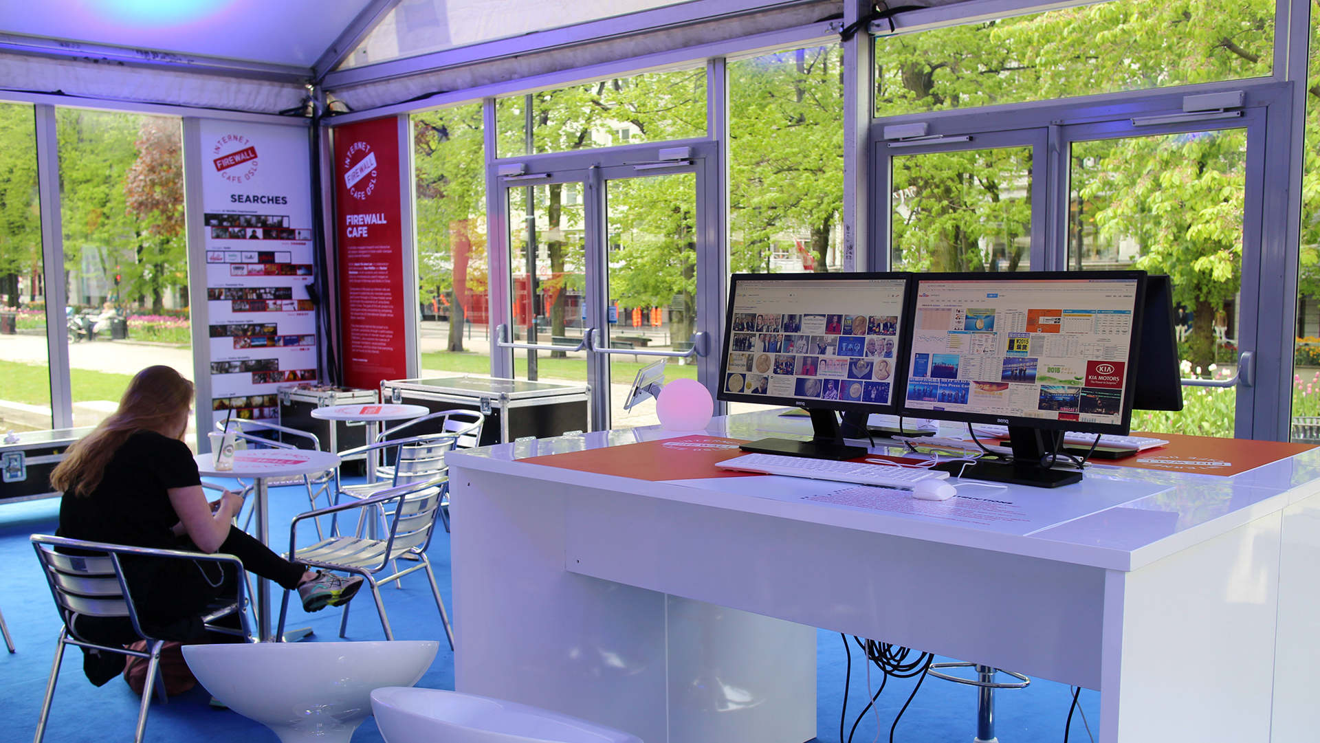 A bright, modern internet café setup featuring a table with two monitors displaying various images and websites. In the foreground, a woman with long hair sits on a chair, using her phone. The café has a blue carpeted floor, surrounded by green trees visible through large windows. There are several white and metallic chairs and tables arranged around the space, contributing to a casual, inviting atmosphere.
