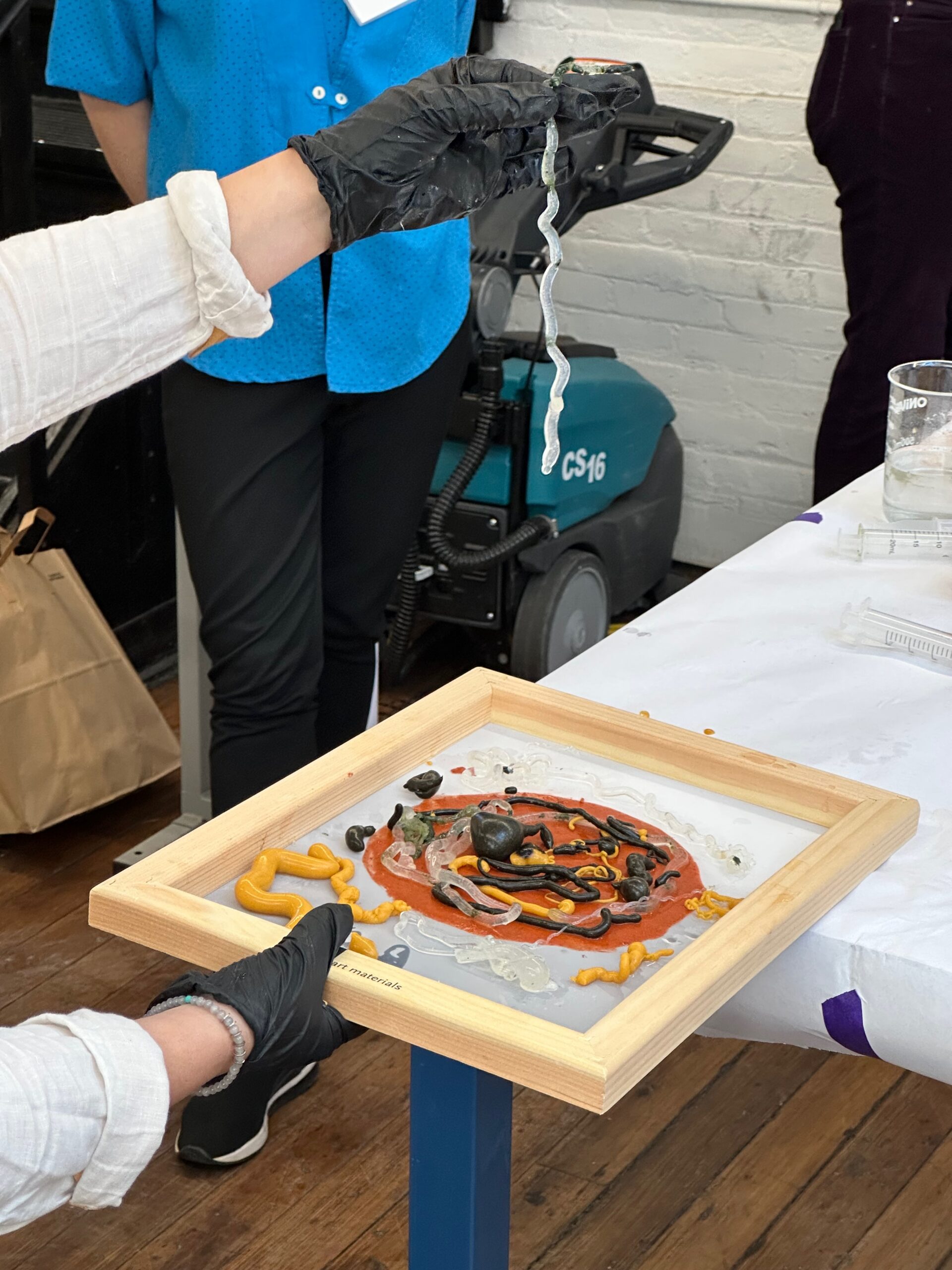 A gloved hand holds a wavy, translucent strand above a framed art piece on a table. The artwork features various colorful materials, including yellow and black strings, arranged on a white surface with a circular orange design in the center. In the background, a person wearing a blue shirt stands nearby, and a cleaning machine is partially visible. The setting appears to be an artist's workshop.