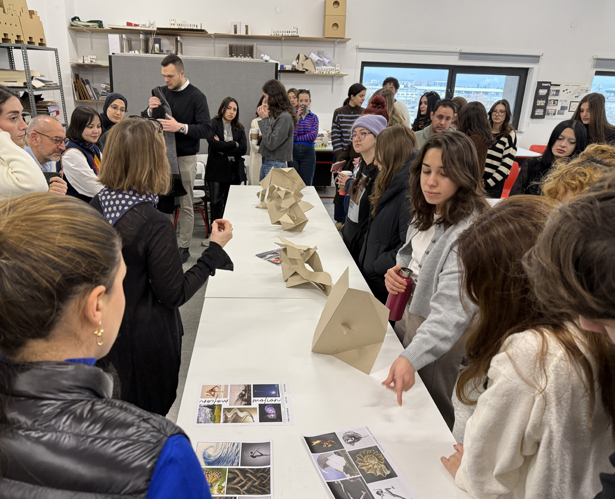 A group of people gathers around a long table exhibiting various folded paper models. Some attendees are engaged in discussion, while others examine visual presentations spread across the table. The setting appears to be a design or architecture workshop, with shelves of materials and models visible in the background. The atmosphere is collaborative and focused on the shared work.