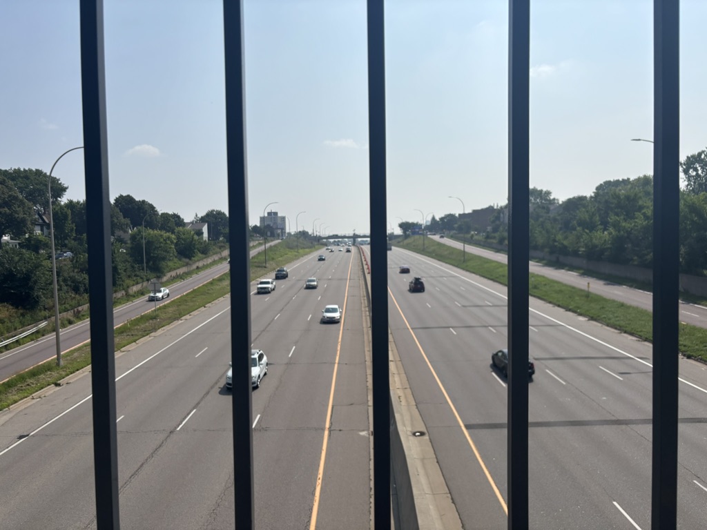 A view of a highway captured from above a railing. Multiple lanes are visible with a few cars driving in both directions. Green trees and buildings line the sides of the highway. The sky is clear with a few clouds. The image suggests a sunny day.