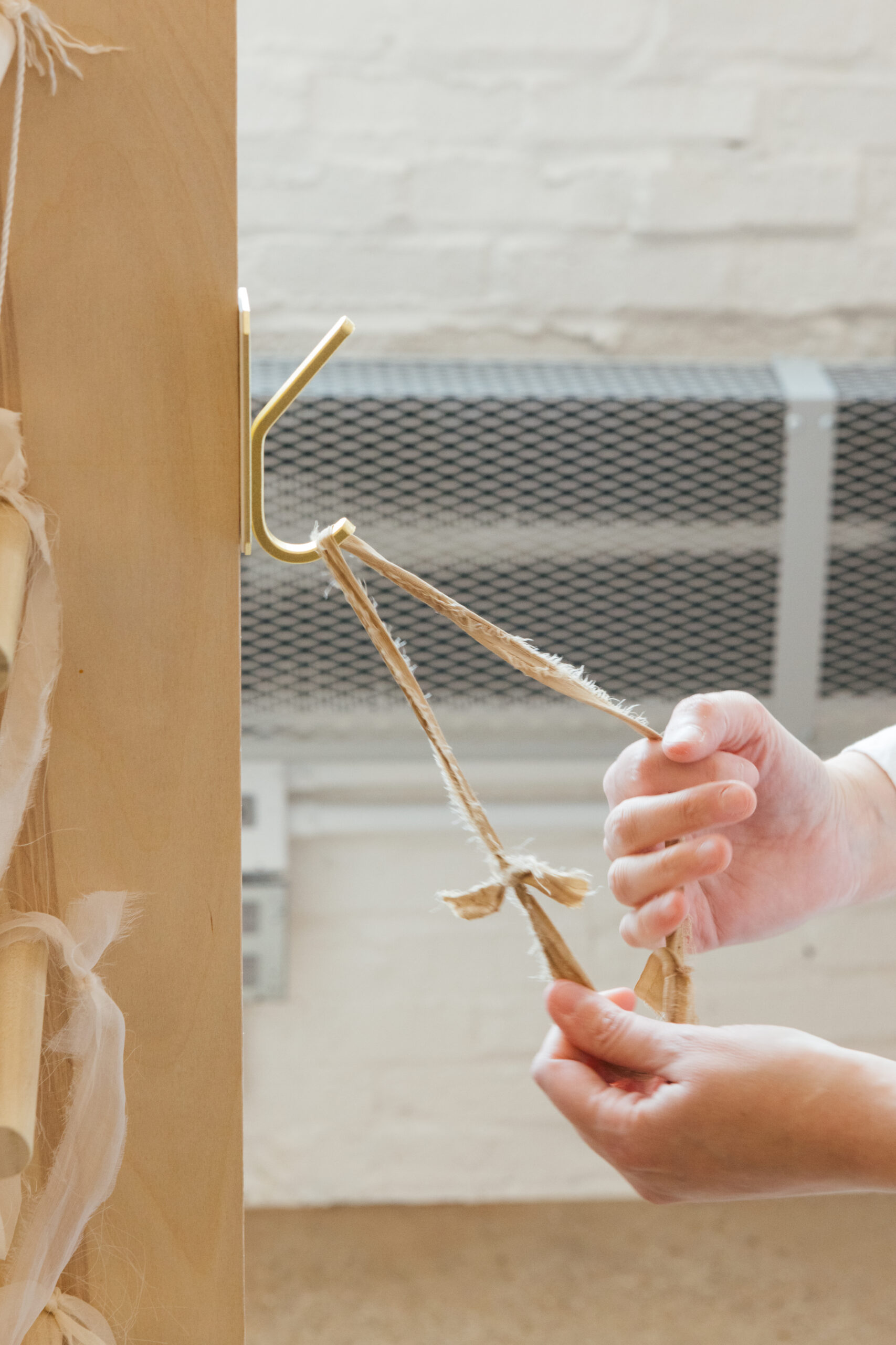 A close-up view of two hands manipulating two pieces of tied fabric or string. The fabric has a natural, textured appearance and is hanging from a gold hook attached to a wooden surface, likely part of a door or wall. In the background, there is a metal grate or vent. The focus is on the hands and the materials being handled.