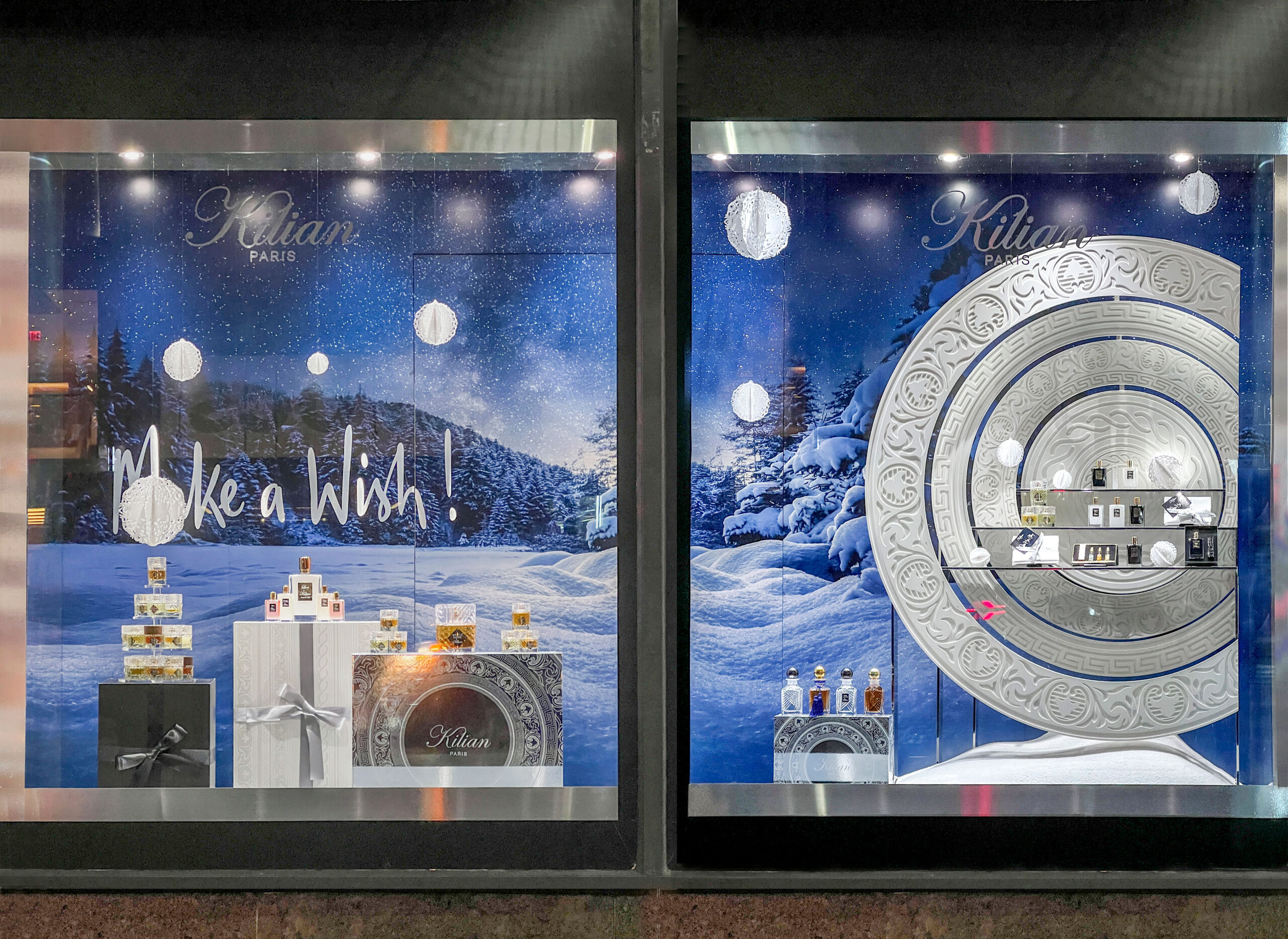 A retail display with two sections featuring holiday-themed decor. The left side has a snowy background with the words "Make a Wish!" and several gift boxes stacked along with perfume bottles. The right side showcases an ornate circular design with various perfume bottles displayed on glass shelves, against a similar snowy landscape backdrop. Soft lights enhance the festive atmosphere, creating a winter wonderland effect.