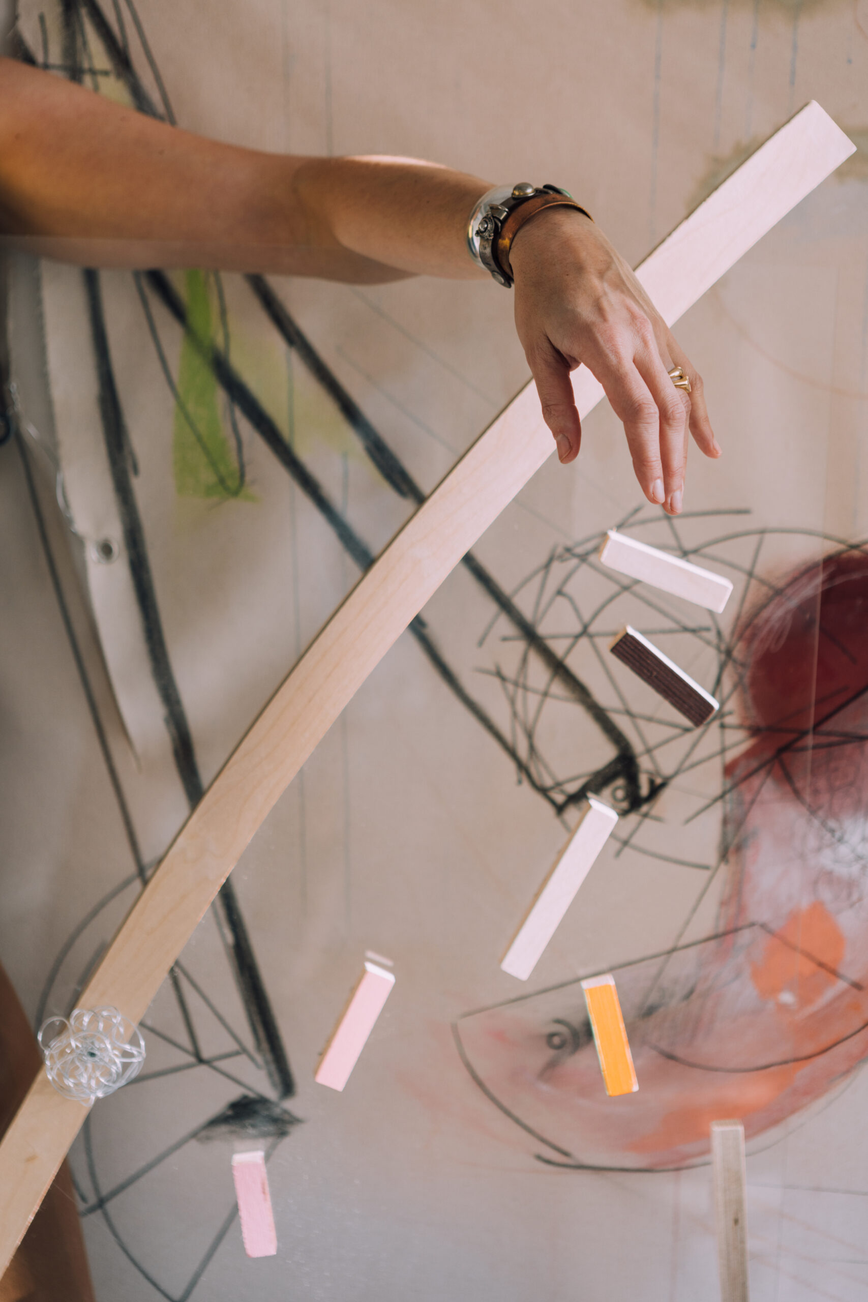 A hand with a silver and brown bracelet is positioned above a transparent structure, which features several colored rectangular pieces hanging in mid-air. The background displays abstract artwork with lines and shapes in various colors, including a prominent red and hints of green. The focus is on the interaction between the hand and the colorful elements.