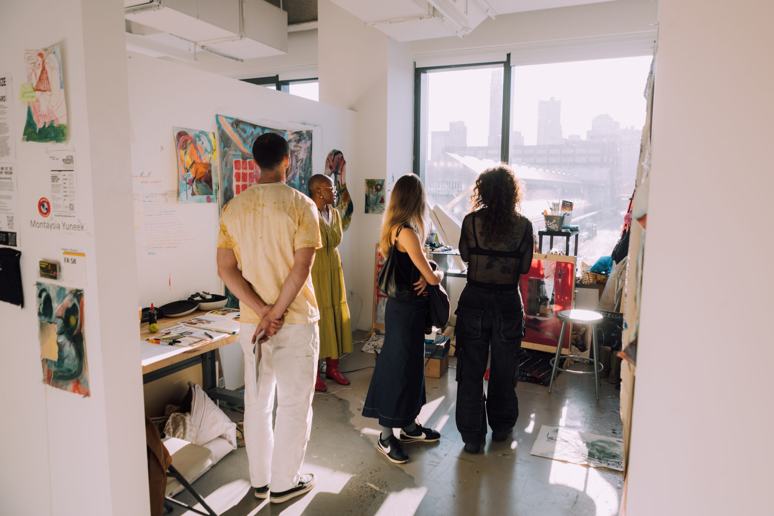 A group of four people stands in an artist's studio with large windows letting in sunlight. They are engaged in conversation, with artworks displayed on the walls. The studio features a table cluttered with art supplies and sketches. One person is wearing a long yellow dress, while others wear casual clothing. The atmosphere appears creative and collaborative.