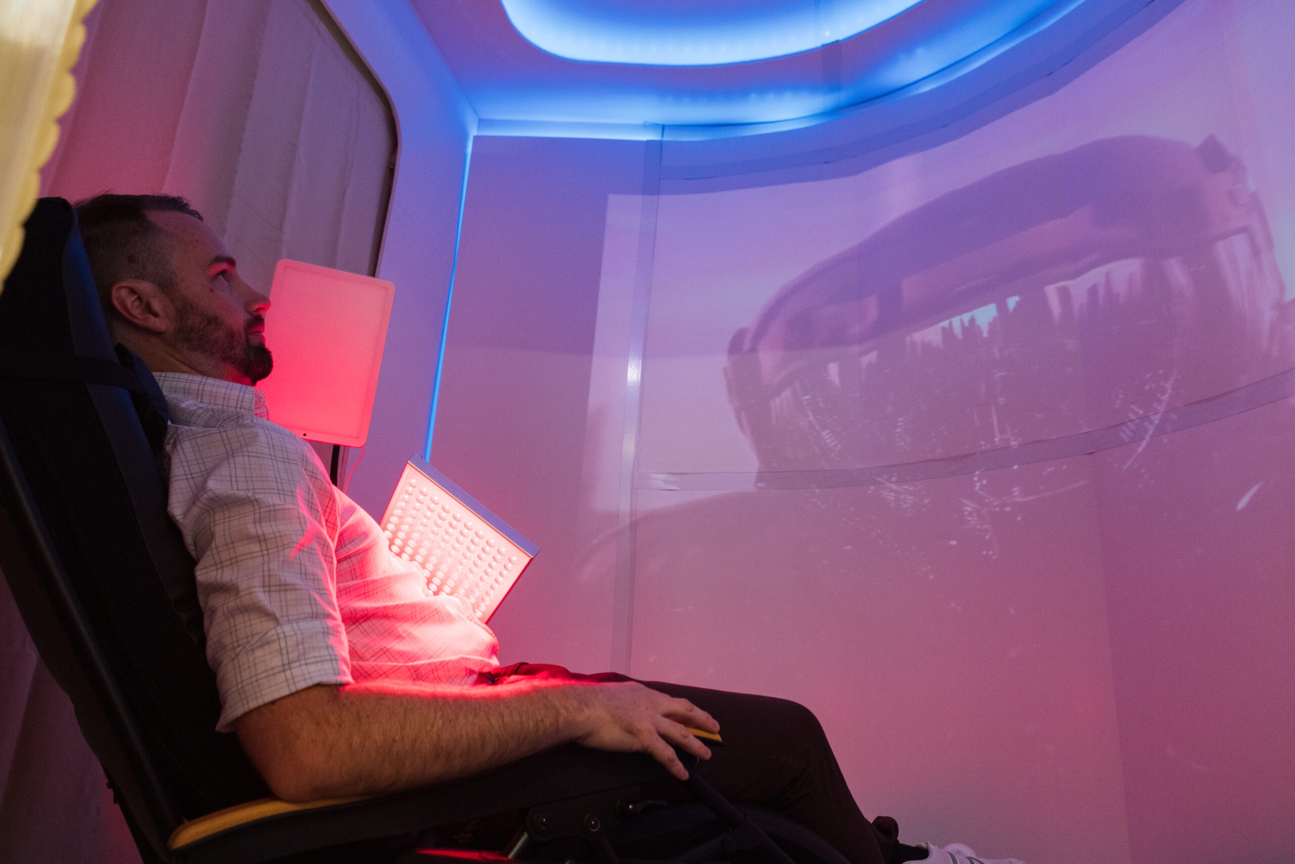 A man is reclined in a chair inside a futuristic pod-like space. Soft lighting in red and blue illuminates the environment. He holds a light panel while gazing at a projection on the curved wall, which appears to show a cityscape. The overall atmosphere suggests relaxation and immersive technology.