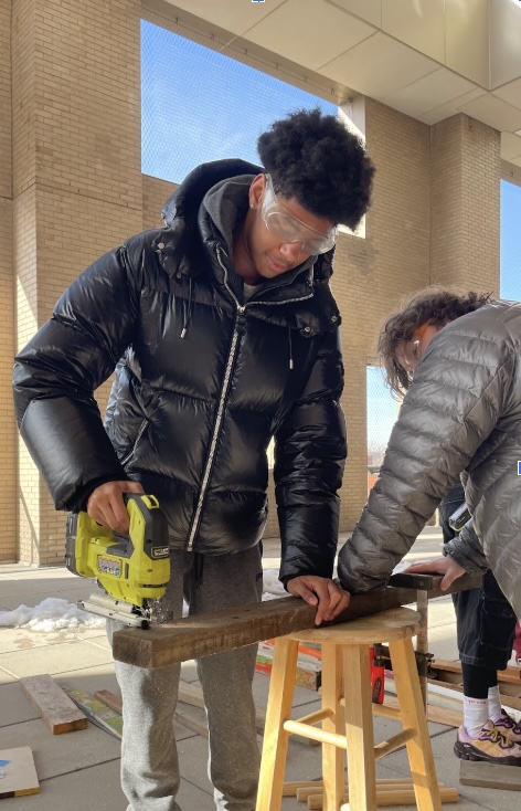 A young person wearing a black puffy jacket and protective goggles is using a jigsaw on a piece of wood while standing at a small stool. Another person, dressed in a gray jacket, is assisting nearby by holding the wood in place. The setting appears to be outdoors with some snow visible on the ground.