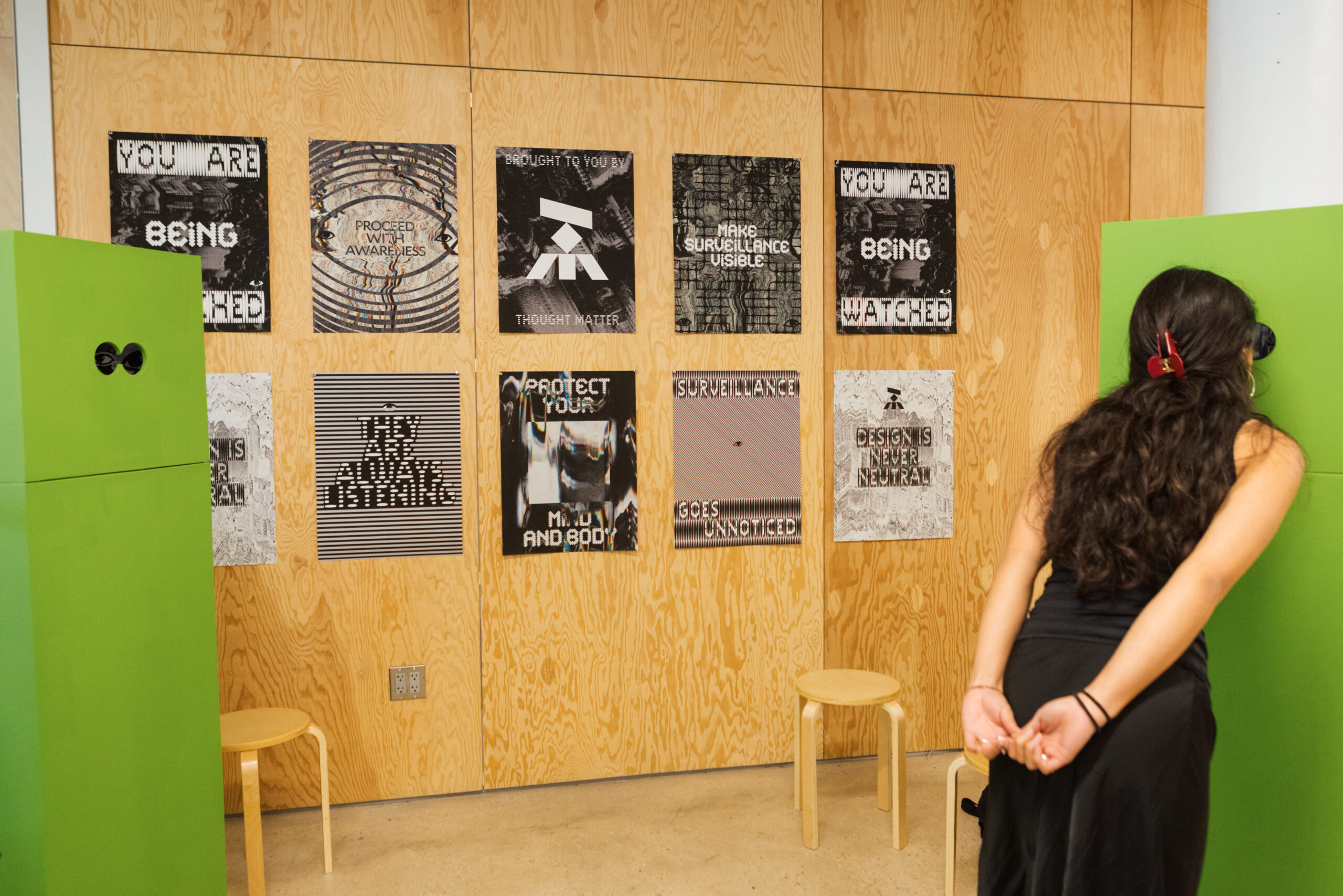 A person with long dark hair stands next to a bright green structure, looking through a circular opening. Behind them is a wall covered with various printed posters that feature phrases related to surveillance, awareness, and protection. Two light-colored stools are visible in the foreground on a smooth floor. The wall is made of light wood, creating a contrast with the colorful elements of the exhibit.