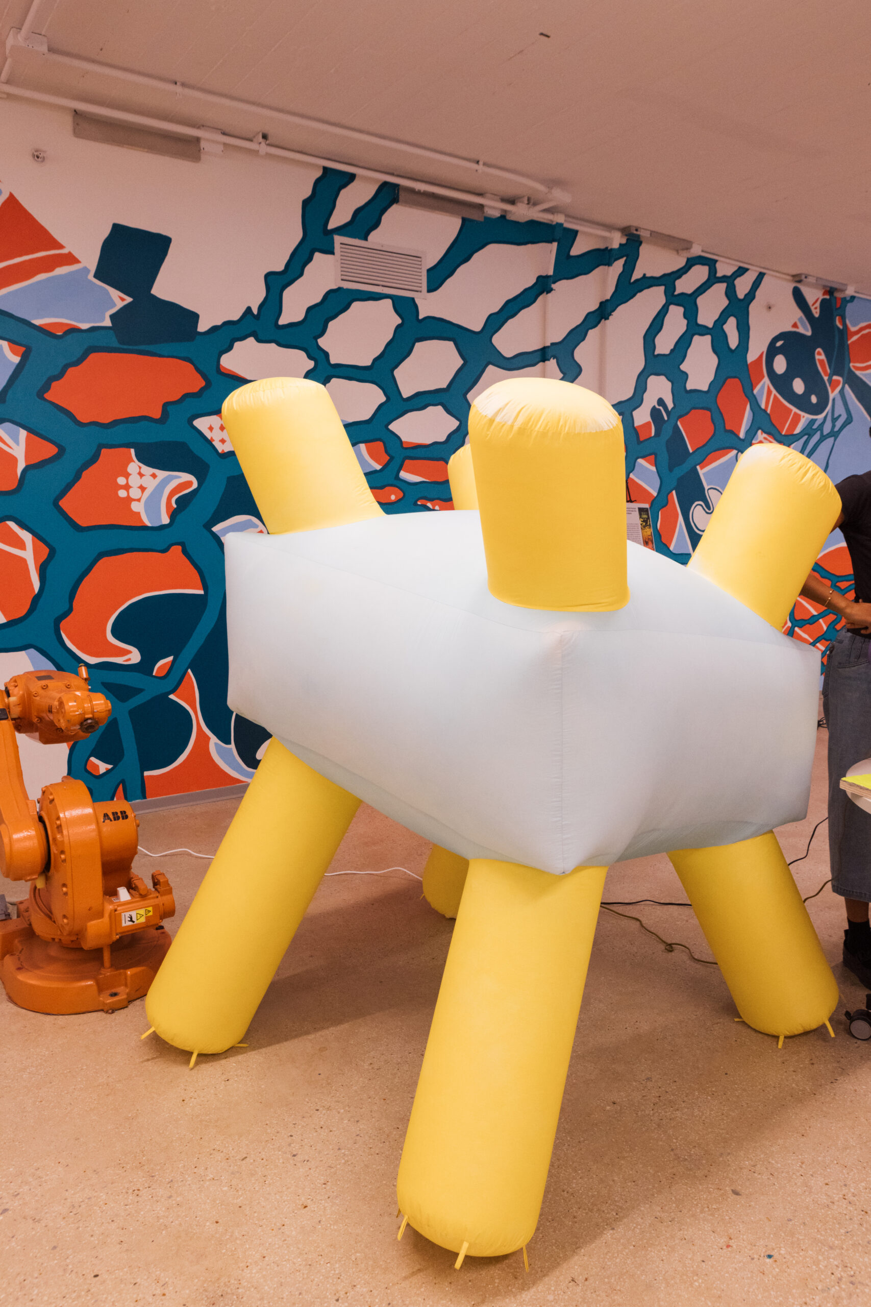 A large, inflatable object with a light blue body and yellow legs is set against a colorful mural featuring abstract shapes in blue, red, and white. An orange robotic arm is positioned nearby. The scene appears to be in a creative or artistic space.