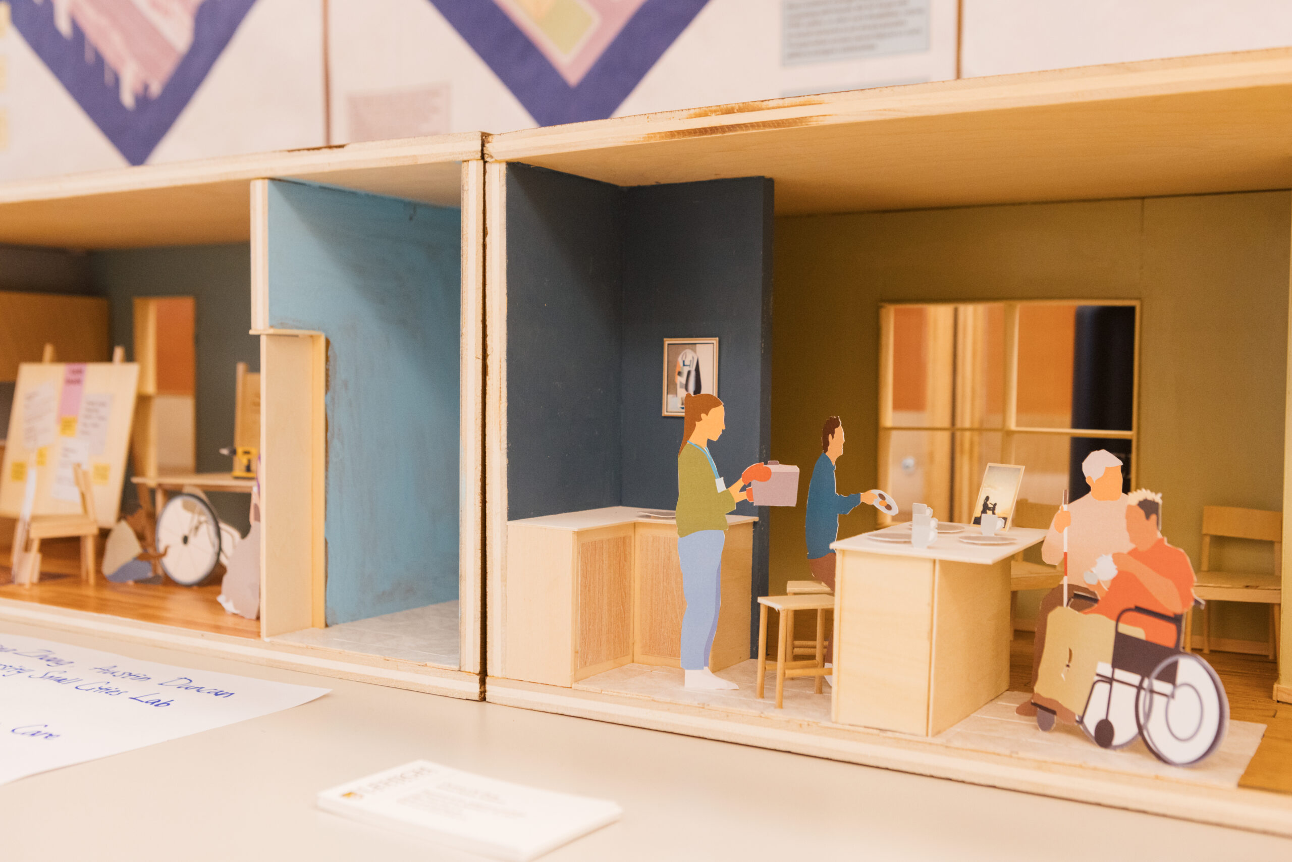 A detailed miniature model of a room is shown, divided into two sections. One side features a figure handing a tray to another person at a table, while another figure stands nearby. The opposite side has an elderly person sitting at a table with a younger individual, accompanied by a wheelchair. Art easels with notes are visible in the background, suggesting an interactive or community space.