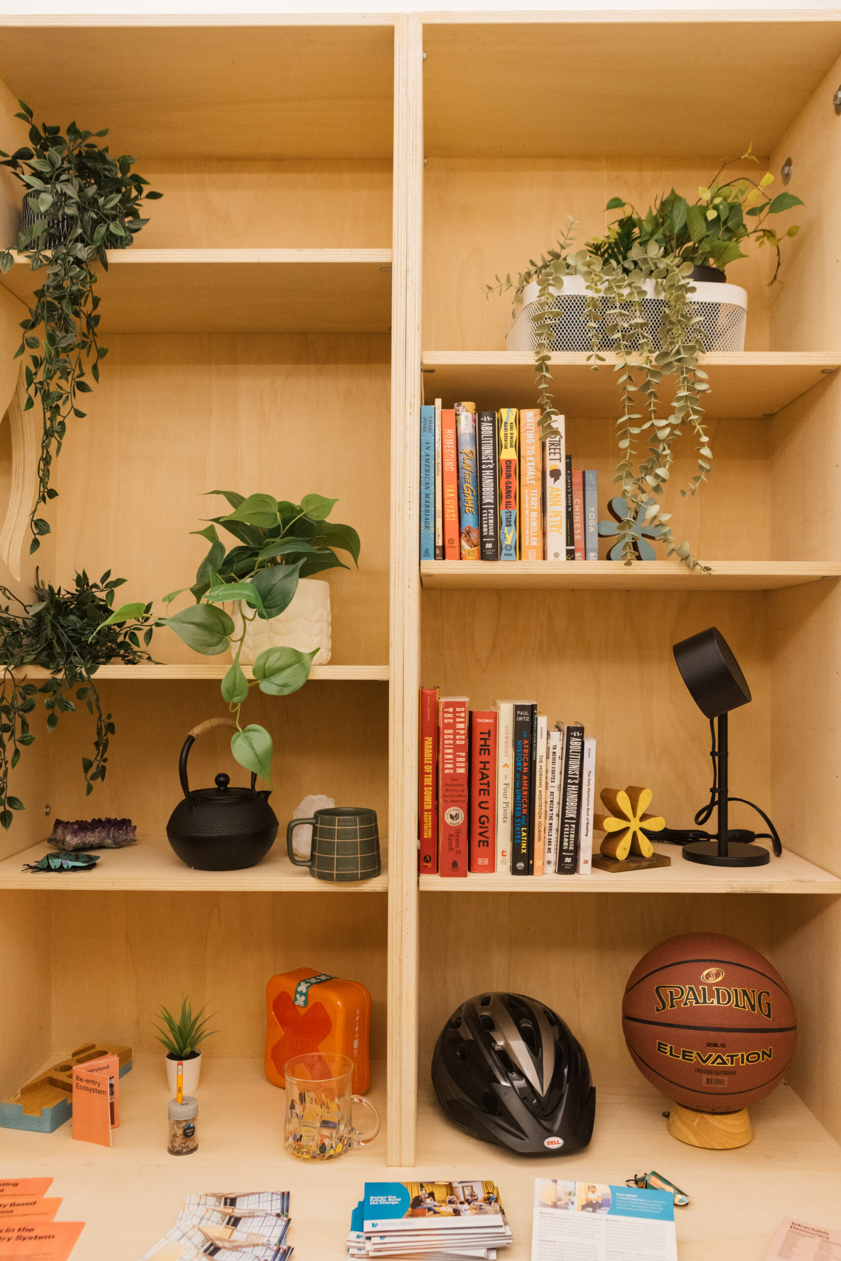 A wooden bookshelf displays books of various colors, decorative plants, and items including a black teapot, a green mug, a small potted plant, and a helmet. A basketball labeled "SPALDING" rests on a wooden stand. The shelf's bottom features a clear glass, a pencil in a jar, and brochures scattered across the surface. A small lamp is positioned on the right side of the shelf.