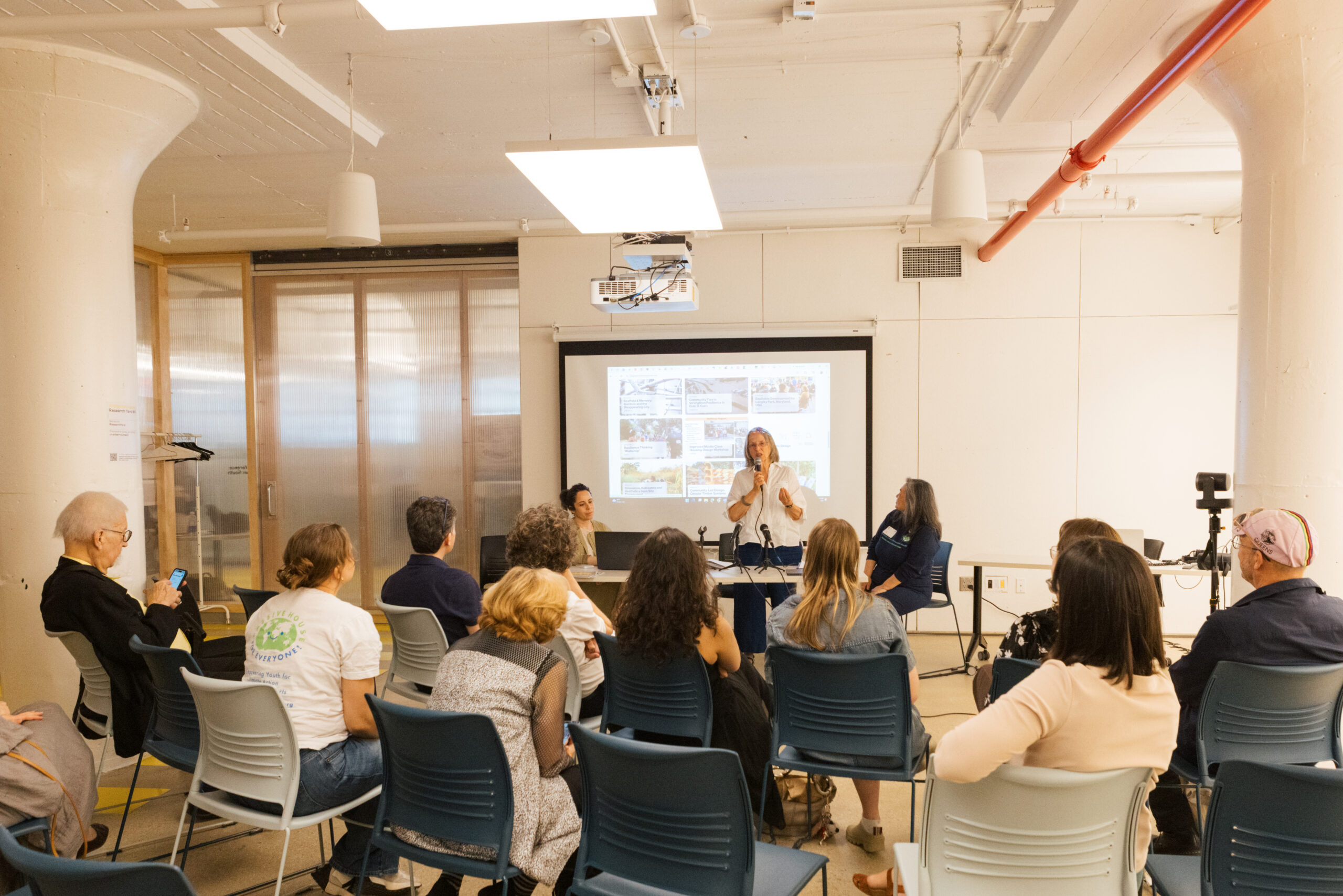 A group of people are seated in a well-lit room, listening to a speaker at the front. The speaker, a woman in a white shirt, is gesturing while talking into a microphone. A presentation is displayed on a screen behind her, showing images and text. The audience includes a diverse mix of individuals engaged in the discussion.
