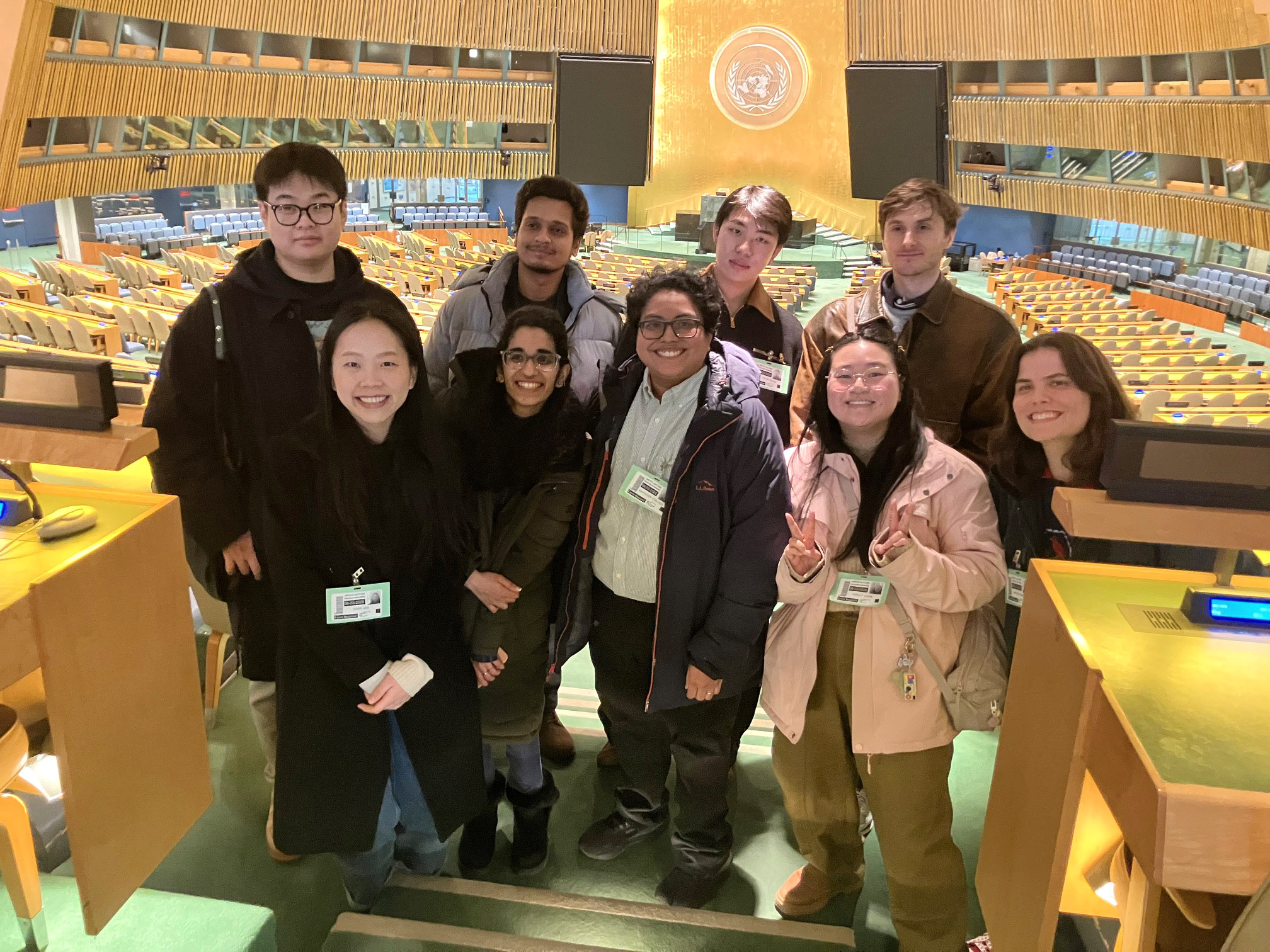A group of eight individuals poses for a photo inside a large hall with empty seats and a prominent United Nations emblem in the background. The group appears to be diverse, wearing casual attire, and they are standing in front of a podium. They smile and display various peace signs, suggesting a friendly and enthusiastic atmosphere. The interior design features wooden accents and a vibrant color scheme.