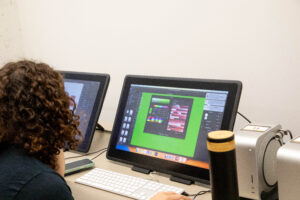 A person with curly hair is sitting at a desk, focused on editing software displayed on a Wacom monitor. The screen shows a digital design with colorful palettes against a green background, while a keyboard and a smartphone are visible on the desk. In the background, another monitor displays a different design. There is also a silver object on the desk that appears to be an audio speaker.