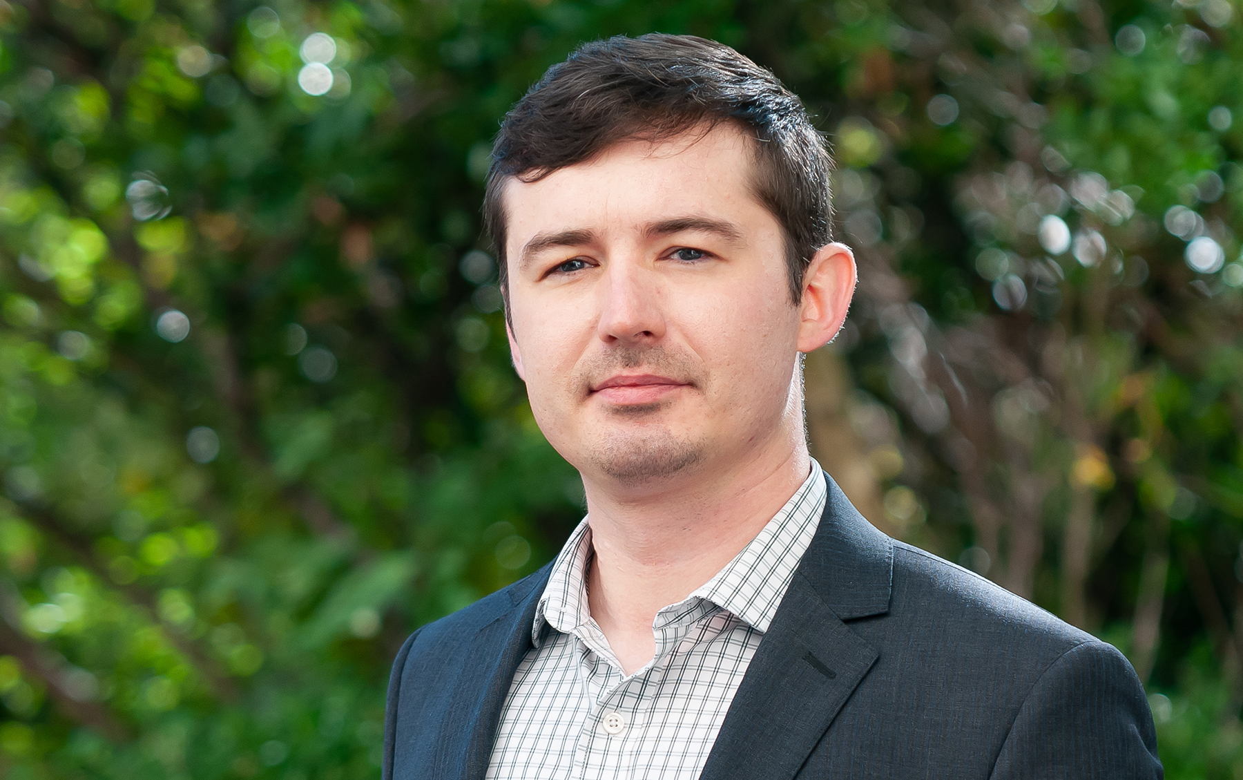 A man with short, dark hair is wearing a gray suit jacket over a light checkered shirt. He has a neutral expression and is standing against a blurred green background filled with foliage.